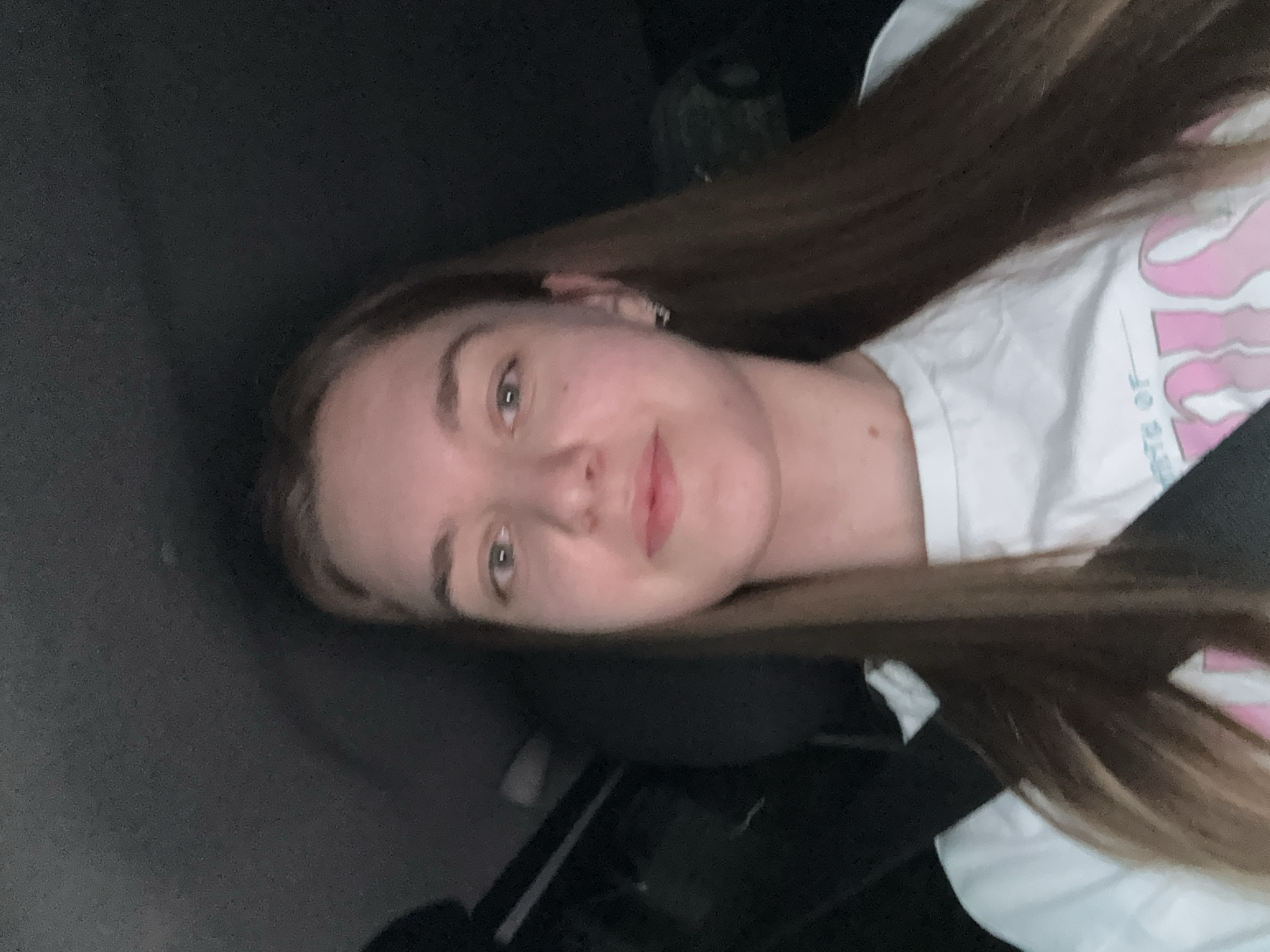 A fair-skinned woman with long brown hair is in a dimly lit interior, possibly a vehicle, wearing a white t-shirt with pink graphic elements. The background appears dark, suggesting nighttime or a low-light environment. The location in the city of Perm, Russia is not evident from the image.