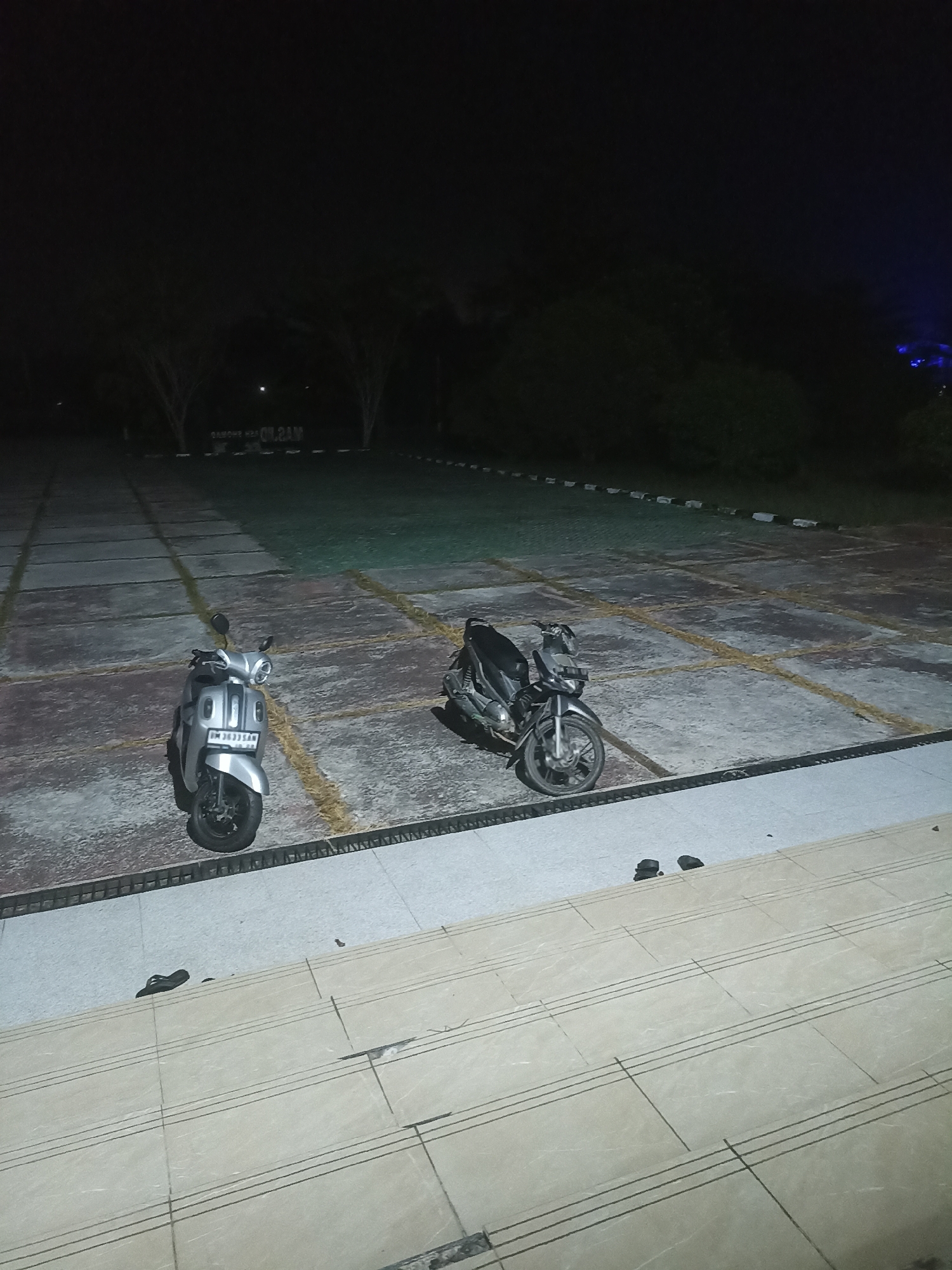The image depicts an outdoor parking lot at night, likely in Sungai Mempura, Indonesia. Two motorcycles are parked on a paved surface marked with yellow lines. One motorcycle, silver in color, is in the foreground on the left, and a darker motorcycle is to its right. In the background, trees are visible, and a sign with white lettering on a dark background is partially illuminated. The ground in the immediate foreground consists of concrete steps with tiled risers. A pair of black flip-flops are present on the steps. The overall lighting suggests artificial illumination from nearby sources.