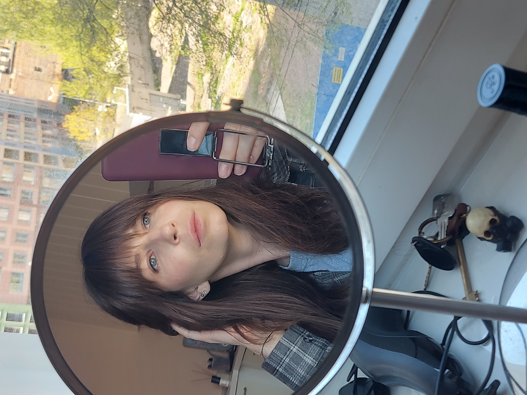 A woman is reflected in a round mirror, holding a burgundy phone. She has brown hair with bangs, blue eyes, and is wearing a gray plaid shirt. The mirror is situated on a white windowsill. A black tube with a white top, keys with a skull keychain, and other miscellaneous objects are also on the windowsill. Outside the window, a park with green trees is visible, as well as buildings indicating a city setting. The city is likely Saint Petersburg, Russia.