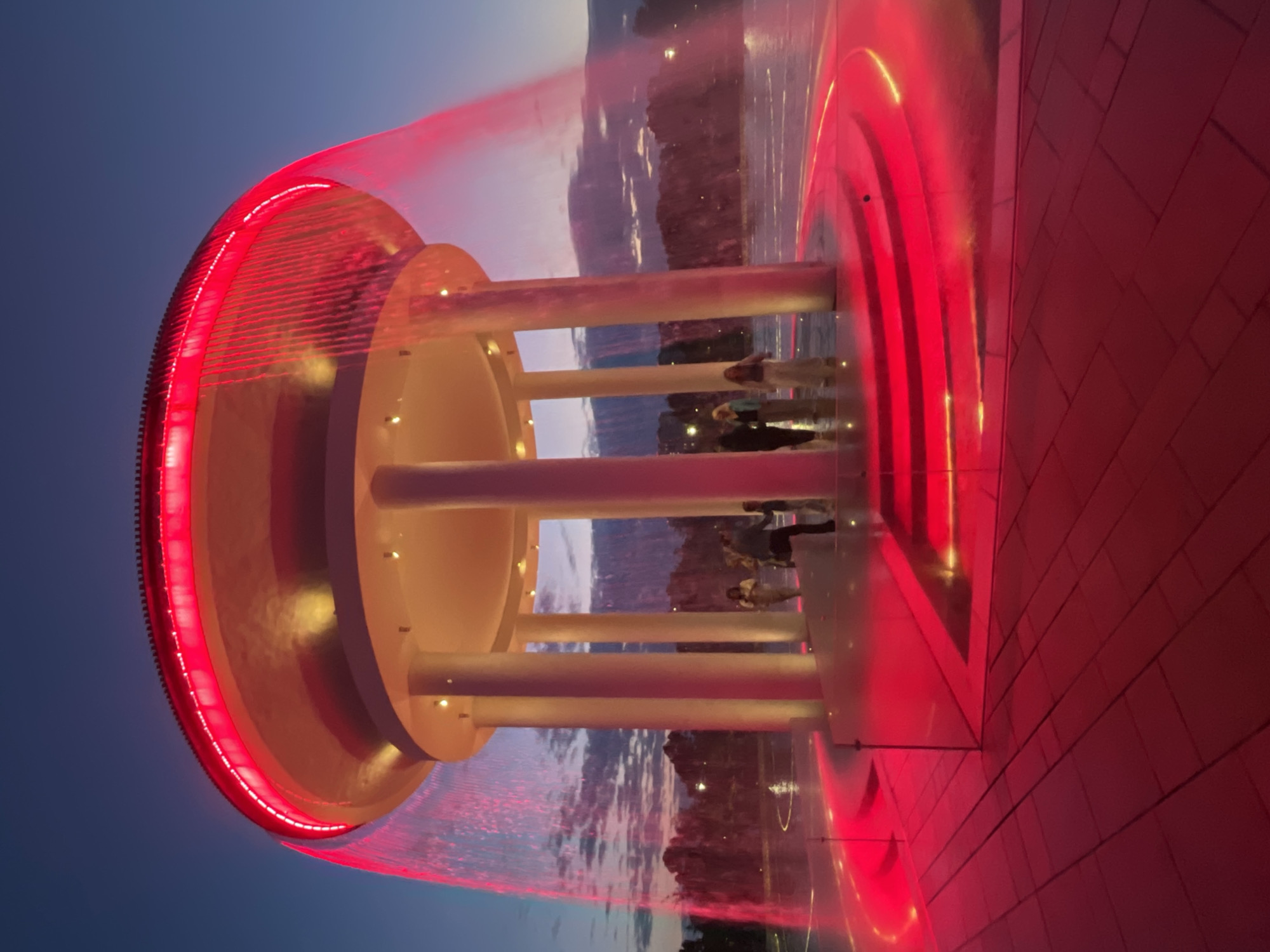 A large, circular, white structure with a domed roof and multiple columns is illuminated by red lights. Water cascades down from the edge of the roof, creating a curtain of red light. The structure is surrounded by a pool of water, also illuminated with red light. Several people are standing on a tiered platform in front of the structure. The sky is dark blue with some clouds, indicating it is dusk or night. The ground in front of the structure is paved with red tiles. This is the "Sun" fountain in Krasnodar, Russia.