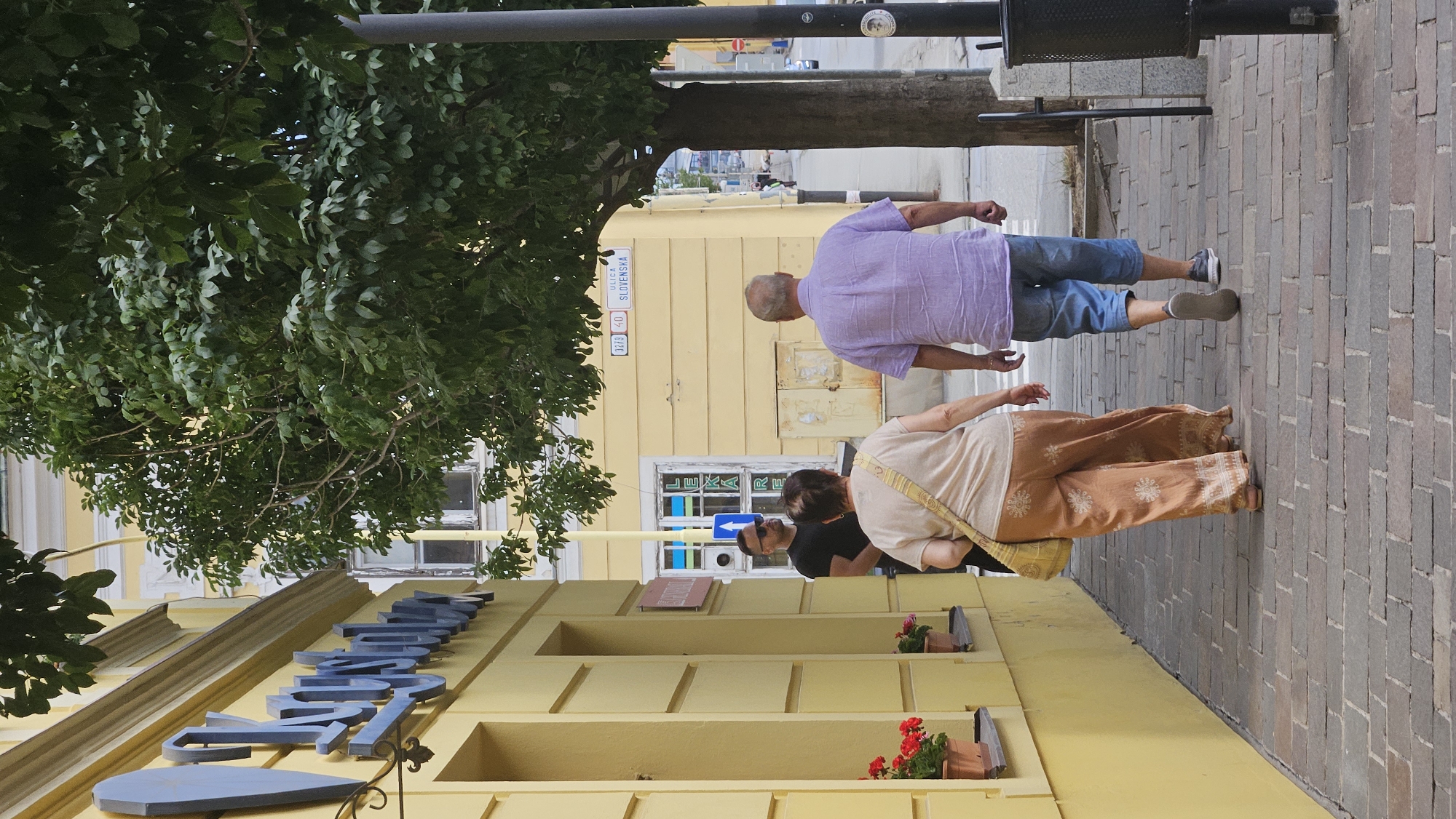 On a cobblestone sidewalk in Prešov, Slovakia, a man wearing sunglasses and a black t-shirt stands near the entrance of a yellow building. A woman with dark hair, wearing a light-colored top and patterned brown pants, walks away from the camera. She carries a yellow bag over her shoulder. Behind her, a man in a light purple short-sleeved shirt and blue shorts walks in the same direction. A tree with green leaves arches over the sidewalk, casting shadows. The building has windows and signage, including blue lettering that appears to spell "Kysuca". A street sign visible on the yellow building reads "ULICA SLOVENSKA". The scene depicts a casual street view with pedestrians and architectural elements.