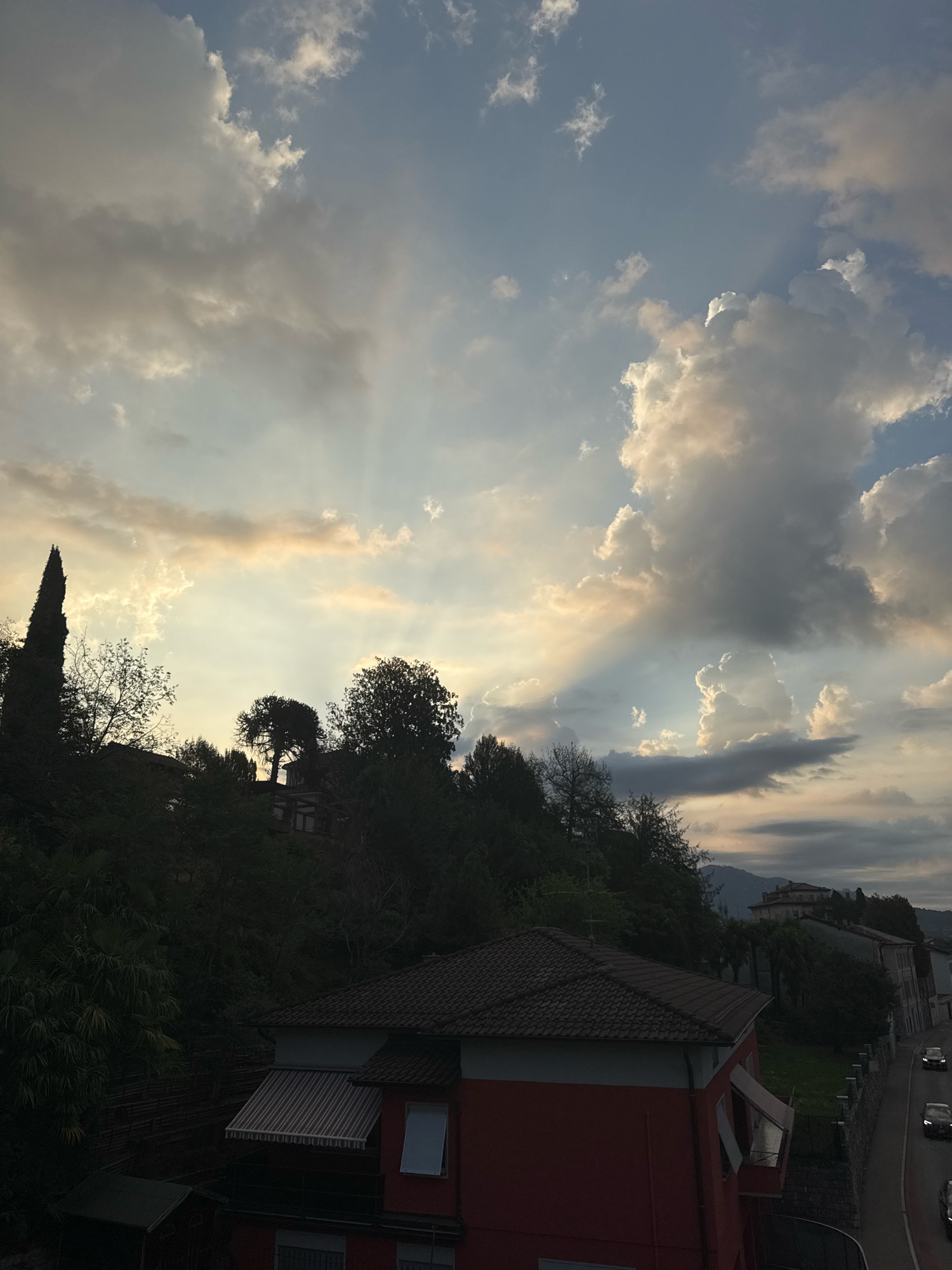 The image depicts a view from a slightly elevated position overlooking a street in Balerna, Switzerland, during a cloudy sunset. In the foreground, a red building with a tiled roof is visible, featuring an awning over a balcony with windows. To the left of the building, lush green foliage and trees dominate the scene, with a tall, dark cypress tree on the left edge of the frame.
The middle ground is characterized by a hillside covered in dense vegetation and trees, with hints of houses or structures nestled within the greenery. The skyline is a dramatic display of clouds illuminated by the setting sun, with visible crepuscular rays beaming through gaps in the clouds. The sky transitions from a pale blue near the horizon to darker grays and whites in the upper portion.
The background shows a road running parallel to the right side of the frame, with several cars in motion. Buildings with pitched roofs are situated along the road and further up the hill. In the far distance, rolling hills are faintly visible against the twilight sky. The overall impression is a serene, residential area at dusk, enhanced by the dramatic cloud formations.