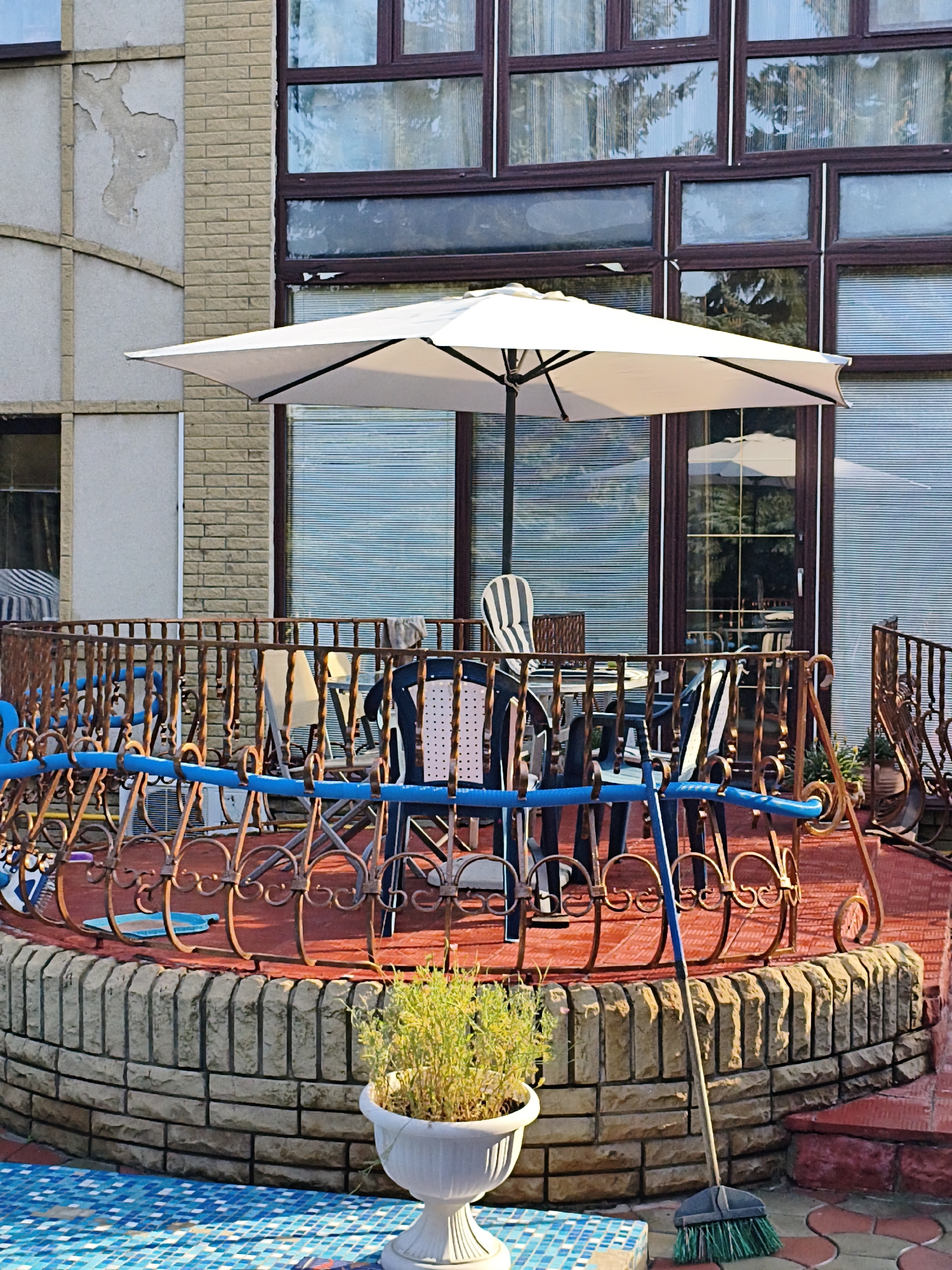 The image depicts an outdoor seating area on a patio with a brick building in the background. A white umbrella is open over a table and chairs. The patio is enclosed by a decorative metal railing. A potted plant sits on a raised brick platform in the foreground, and a broom leans against the wall. The flooring is a mix of red tiles and a blue mosaic pattern. The building has large windows with blinds. This scene is located in Makiivka, Ukraine.