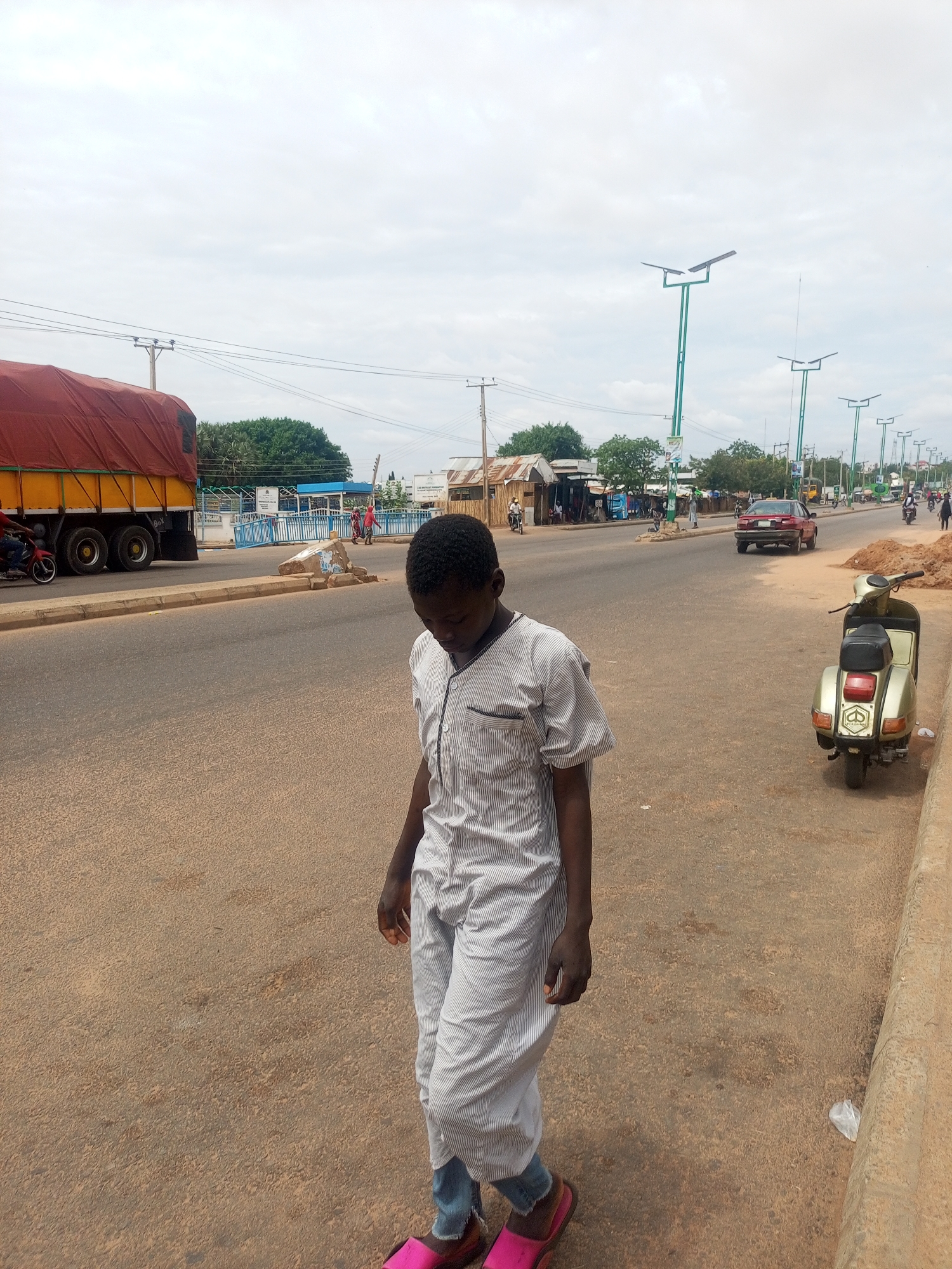 The scene depicts a street in an unknown city and country, observed on an overcast day. A young individual with dark skin and short dark hair, wearing a light-colored striped tunic over light blue trousers and bright pink slide sandals, walks in the foreground on the right side of the paved road, looking downwards.
The multi-lane road, characterized by brownish-grey asphalt, extends into the distance. On the left side of the road, a large truck with a red tarp covering a yellow cargo bed is partially visible, appearing to be in motion. A person on a red motorcycle is also visible near the truck. In the middle ground, a red sedan is driving away from the viewer.
On the right side of the road, beyond the foreground subject, a vintage-style light green or gold scooter with a black seat is parked on a dirt verge next to a concrete curb. A pile of reddish-brown dirt and a white plastic bag are on the ground near the scooter.
The street is lined with numerous tall, green streetlights featuring solar panels, stretching into the background. Traditional utility poles with overhead power lines are also visible against the cloudy sky. In the mid-ground, a concrete barrier or block separates lanes, behind which blue railings and several distant figures are discernible. Along both sides of the road in the background, various single-story buildings with corrugated metal roofs are present, interspersed with green trees and bushes. Several individuals are seen walking or riding motorcycles further down the street, indicating activity in the area.