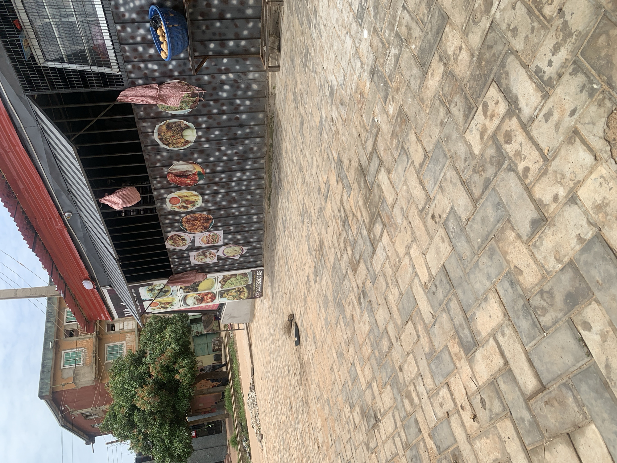 The image depicts a street scene in Dadi Riba, Nigeria. In the foreground, a paved courtyard made of interlocking grey and tan bricks is visible. A broom and a flat object, possibly a shoe, lie on the ground. To the right of the courtyard is a building with a red corrugated metal roof and a dark metal wall adorned with circular pictures of food. A blue plastic basket filled with potatoes sits on a shelf attached to the wall. Further left, a multi-story building with orange and light brown facades stands behind a tree with green foliage. A few individuals are casually standing or sitting near the base of the building. The sky is overcast.