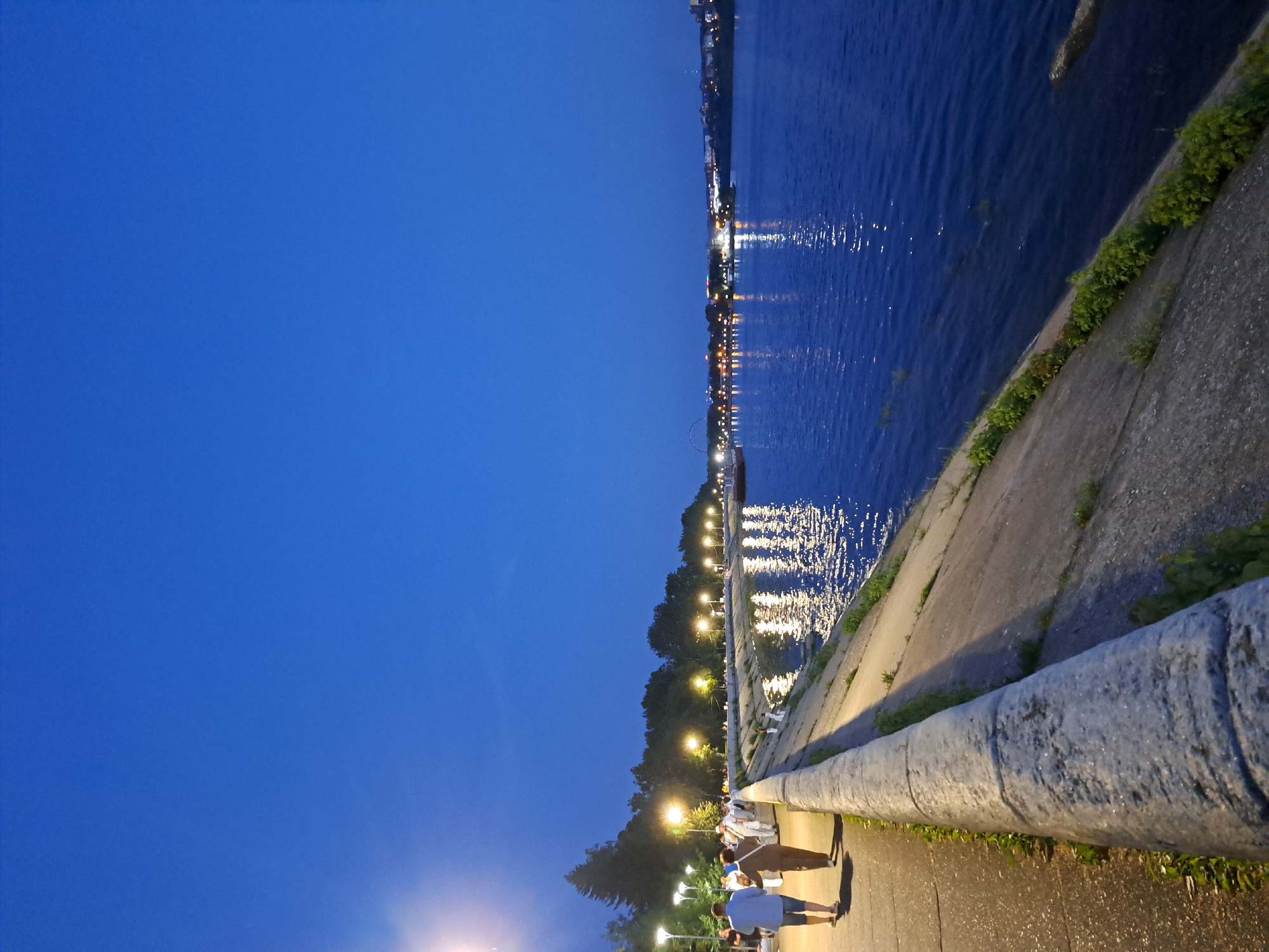 This image depicts the Angara River embankment in Irkutsk, Russia, during the evening. The sky is a deep, clear blue, suggesting twilight. The embankment itself is constructed of concrete and stone, with a paved walkway running alongside it. Several lampposts are visible, casting pools of light on the walkway and illuminating trees and foliage lining the riverbank.
On the walkway, two figures are walking away from the camera, a man in a light blue shirt and shorts and a woman in a brown top and light-colored pants. Other figures are also visible further down the walkway, some appearing to be walking in groups. The river is a dark blue, reflecting the lights from the opposite bank, which is dotted with buildings and more lights. The overall atmosphere is peaceful and urban.
