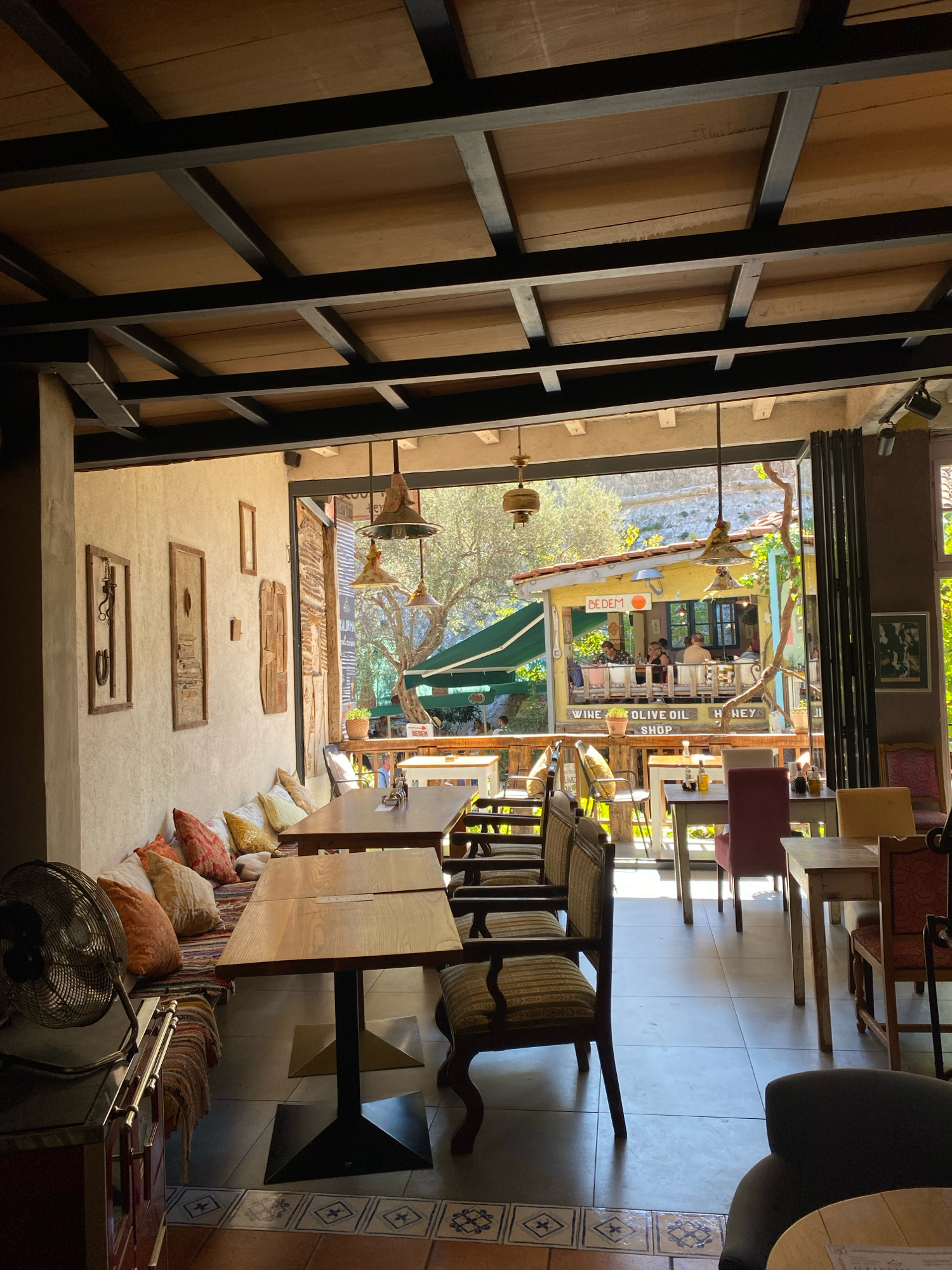 This image depicts the interior of a restaurant or cafe located in Stari Bar, Montenegro, with a view extending to an outdoor seating area. The interior features a rustic design with a wooden slatted ceiling supported by dark metal beams. One wall is adorned with framed artwork and decorative pieces, and a long, cushioned bench with numerous colorful pillows runs along it. Several wooden tables and chairs are arranged throughout the indoor space.
A large opening, possibly a folding door or window, leads to an outdoor area. This outdoor section is partially covered by a green awning and features more tables and chairs. In the background, a small yellow building with the sign "BEDEM" is visible, suggesting it's part of the establishment or a neighboring business. People are seated at tables in both the indoor and outdoor areas, some engaged in conversation and eating. Natural sunlight streams into the interior, creating distinct shadows. The flooring in the foreground consists of large grey tiles with a decorative border. A vintage-style fan is positioned in the lower left corner of the image. The overall ambiance suggests a relaxed dining environment in a Mediterranean setting.
