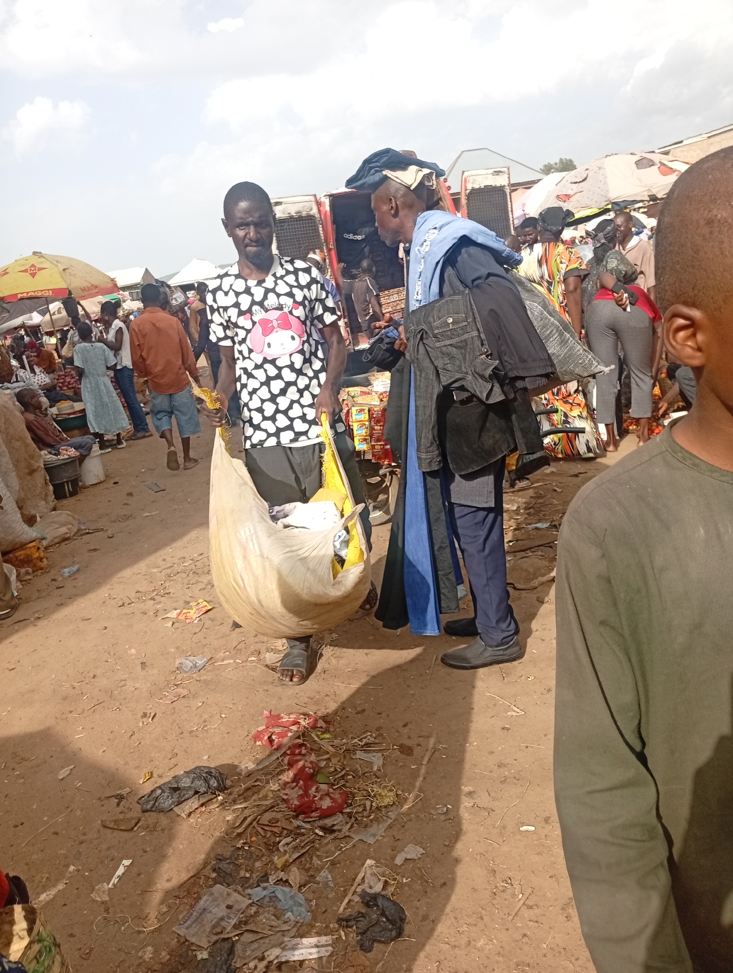 The image depicts a bustling outdoor market scene in Rafin Par, Nigeria, likely during the daytime given the bright sunlight casting shadows. The ground is dusty and littered with debris, suggesting a busy, perhaps unpaved, area. In the foreground, a man wearing a black and white patterned t-shirt with a cartoon character and dark pants, is carrying a large, light-colored sack over his shoulder. Next to him, another man, dressed in dark pants and a blue top with clothes draped over his arm, stands looking to the side.
In the background, more people are visible, engaged in various market activities. Some appear to be browsing goods, while others are interacting. Market stalls and umbrellas, including one with "MAGGI" visible, suggest the sale of various products. The overall atmosphere seems lively and active, typical of a local marketplace. The weather appears to be clear and sunny, with some clouds in the sky. The scene is characterized by a mix of individuals and the general commercial activity of a Nigerian market.
