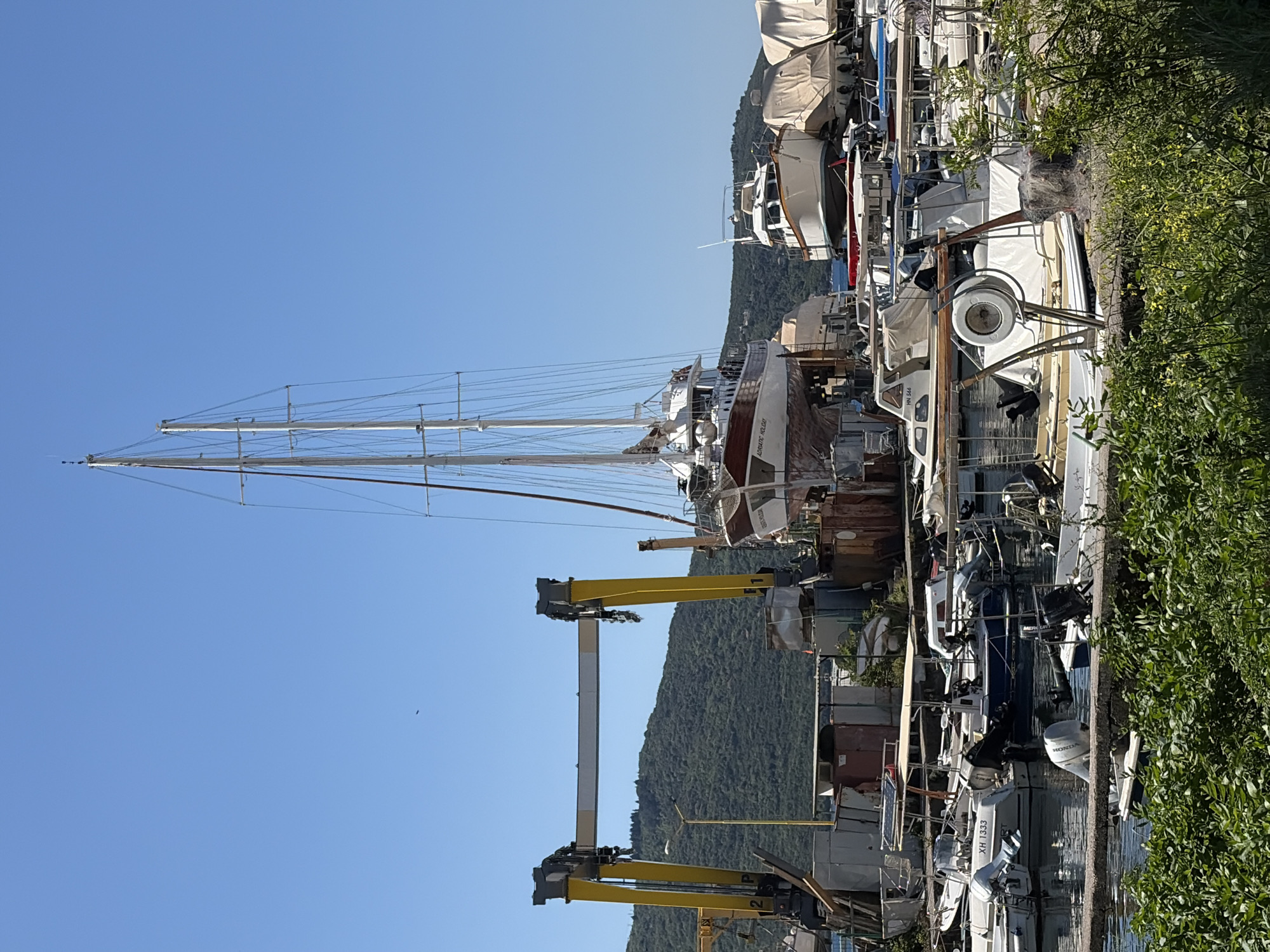 The image shows a boatyard in Zelenika, Montenegro. Several boats are docked and stored on land, including sailboats and smaller motorboats. A large crane is visible, likely used for moving boats. The scene is set against a backdrop of a clear blue sky and a green, tree-covered hill. The location appears to be a working boatyard, with evidence of maintenance and storage activities. Vegetation is present along the edge of the scene, adding a natural element to the industrial setting.