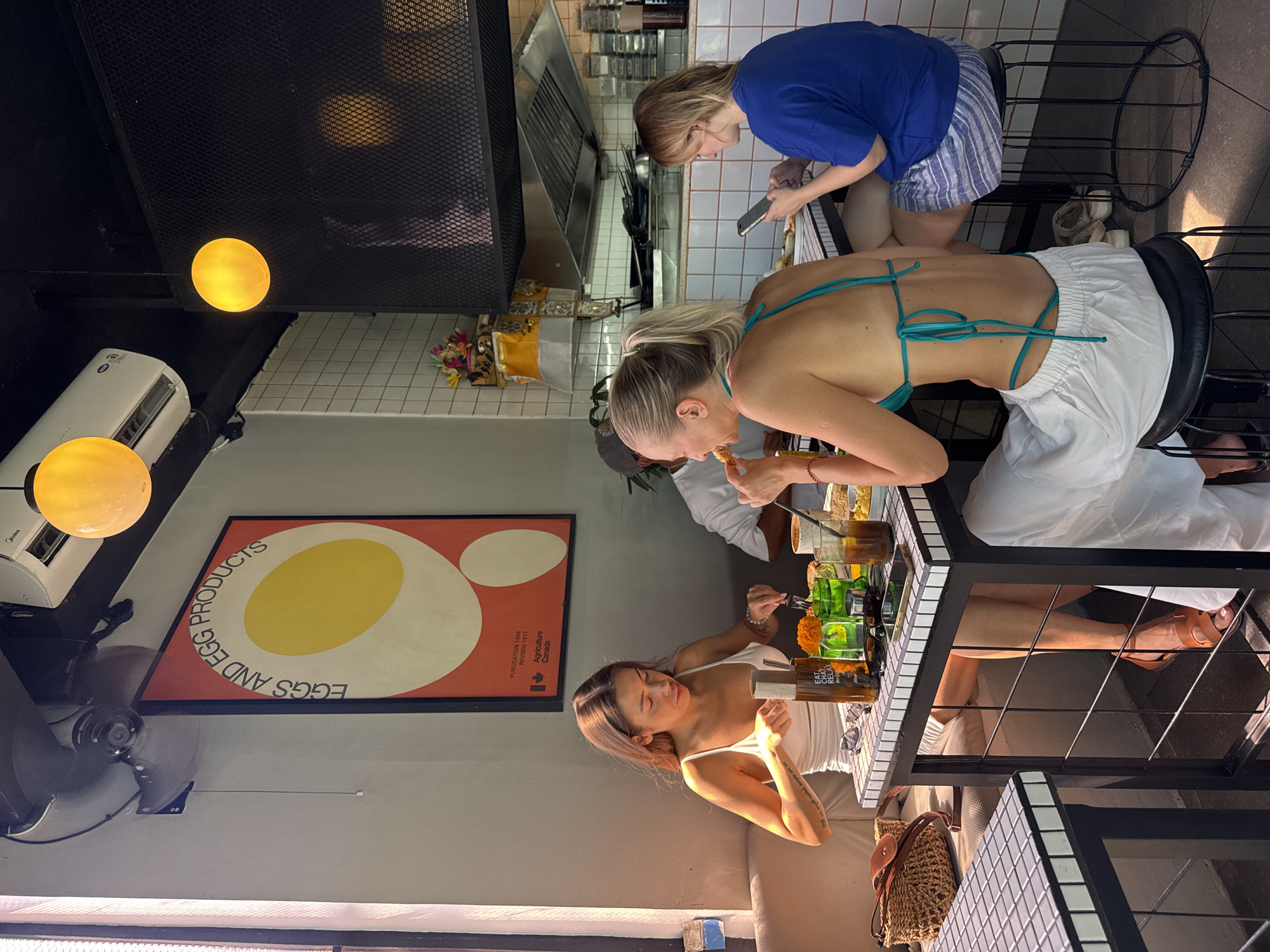 Three women are sitting at a table in a restaurant, engaged in eating and using mobile devices. The woman closest to the camera wears a white top and has visible tattoos on her arm. The woman in the middle is wearing a turquoise bikini top and white pants. The third woman is wearing a blue top and striped shorts, and is looking at her phone. On the table are various beverages and food items. The restaurant has a modern design with black and white tile tables. The restaurant is located in North Kuta, Indonesia as indicated by the text visible on a bottle on the table. A poster on the wall features a graphic of eggs with text "EGGS AND EGG PRODUCTS." An air conditioning unit and a hanging fan are visible on the ceiling.