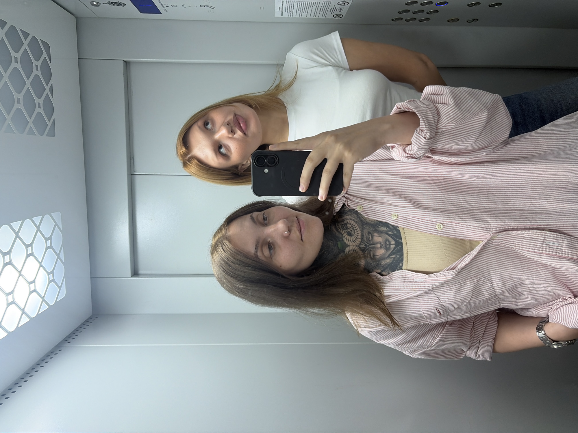 Two individuals are posing for a selfie in an elevator. The person in the foreground, on the left, has long brown hair and is wearing a light pink and white striped button-up shirt over a beige tank top. They have a prominent tattoo on their neck and chest. The person in the background, on the right, has blonde hair and is wearing a white t-shirt. They are looking upwards. The elevator has gray walls and a white ceiling with a patterned light fixture. An elevator control panel with illuminated buttons is visible on the right side of the frame. The image is a selfie taken with a smartphone held by the person on the left. The location is the city of Chaikovsky, Russia.
