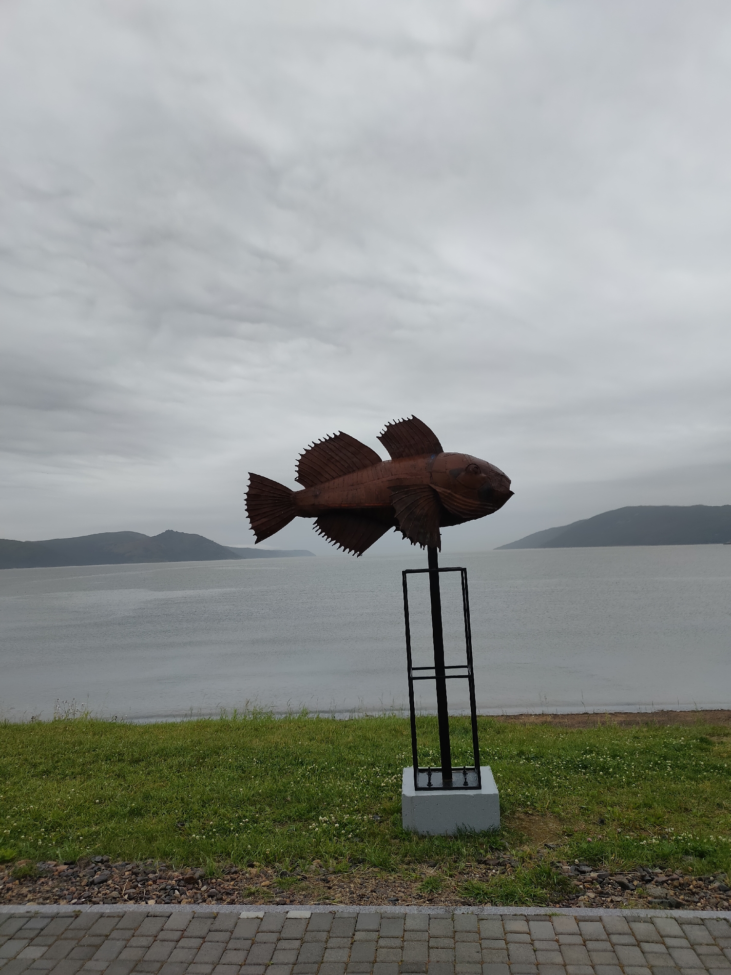 A metallic sculpture of a fish is mounted on a black metal stand atop a concrete base. The sculpture, appearing to be made of weathered copper or bronze, depicts a fish with detailed fins and scales, facing to the right. The sculpture is positioned in a grassy area adjacent to a paved walkway. In the background, a calm body of water stretches towards distant, muted green hills under an overcast, cloudy sky. The overall scene suggests an outdoor public space, potentially a waterfront park or promenade in Magadan, Russia, given the landscape features. There are no people or animals visible in the image.