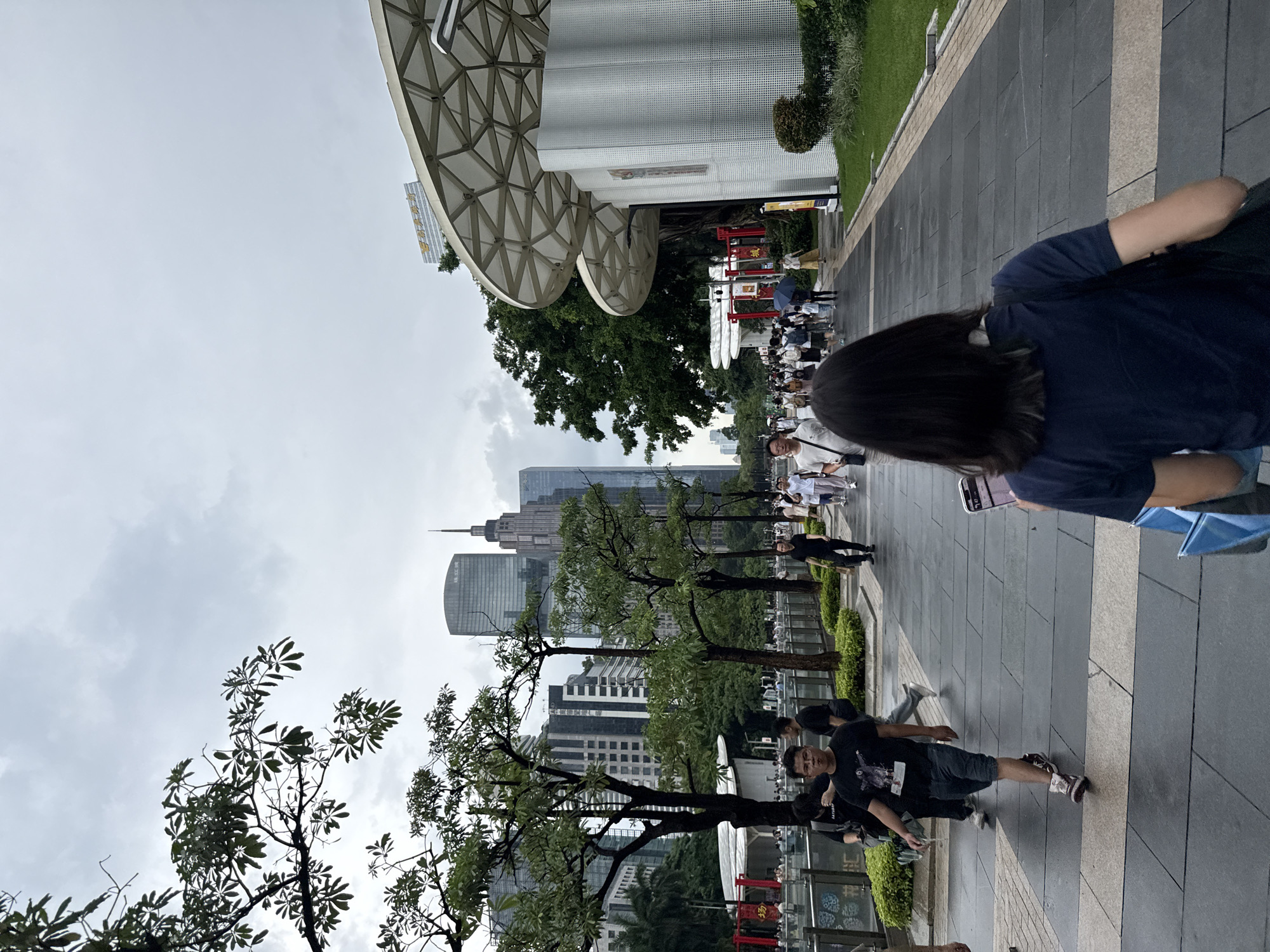The image depicts a street scene in Tianhe District, China, on a cloudy day. A group of people are walking along a paved walkway lined with trees and buildings. The architecture of the buildings suggests a modern urban environment, with some featuring geometric patterns on their exteriors. In the foreground, a person is seen from behind, wearing a dark blue t-shirt and carrying a bag. Further down the walkway, other individuals are visible, some holding umbrellas, indicating that it might be raining or has recently rained. The overall atmosphere appears to be casual, with people going about their day. The presence of trees and greenery amidst the urban landscape suggests a well-maintained public space. The lighting is subdued due to the overcast sky, suggesting it is daytime, possibly morning or afternoon.