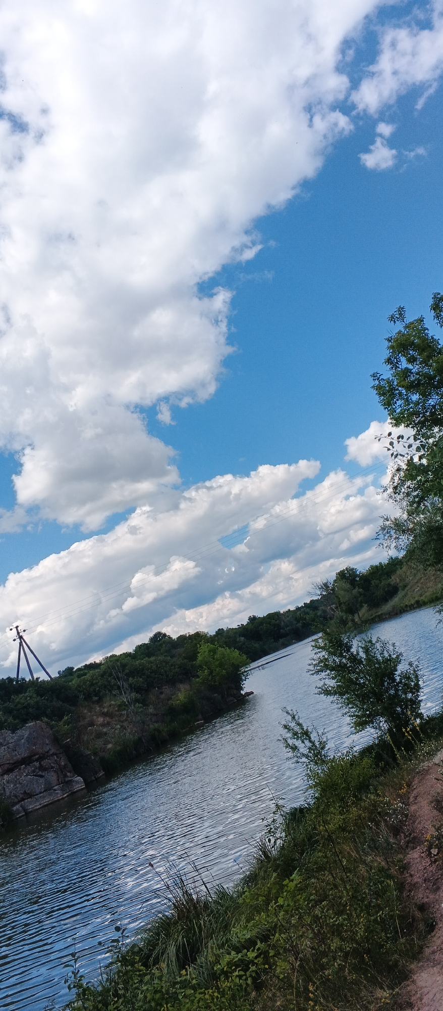 The image depicts a body of water with lush green vegetation along its banks, under a bright blue sky dotted with fluffy white clouds. A rocky outcrop is visible on the left side of the frame, near a wooden power pole with insulators. The water is calm, reflecting the sky and surrounding trees. On the right bank, there are more trees and some dry, dirt ground. The perspective is from a slightly elevated position, looking across the water towards the tree-lined horizon. The scene suggests a natural, perhaps semi-rural, setting.