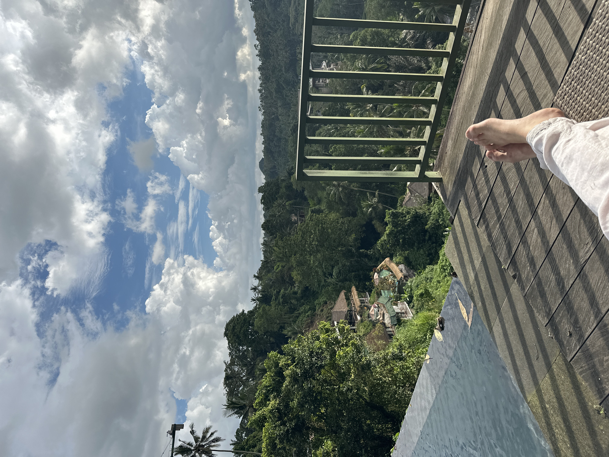 The image depicts a scenic view in Ubud District, Indonesia, featuring a person relaxing on a wooden deck overlooking a lush, dense forest. A green metal railing separates the deck from the verdant landscape. Parts of buildings are visible nestled amongst the trees. In the foreground, a portion of what appears to be a pool is visible. The sky is filled with a mix of blue patches and cumulus clouds. A pair of bare feet are visible resting on the wooden deck. The perspective suggests the photo was taken from a high vantage point.