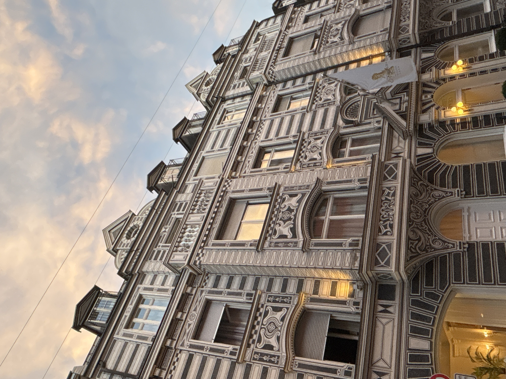 The image shows the facade of a building in Munich, Germany, featuring intricate decorative patterns. The building has multiple stories with various windows, some with shutters. A flag with a crest is visible on the facade. The sky above has a cloudy, sunset or sunrise appearance. The architectural style suggests it is likely a historic building in a city center. The lighting indicates it is either early morning or late evening.