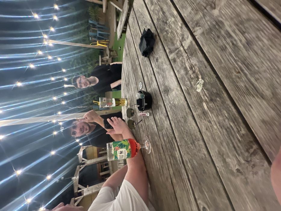 Three people sit at a wooden table under string lights. A glass of beverage with tropical print, another taller glass with beer, sunglasses, an ashtray and a pair of black gloves occupy the table. One person in a black shirt is taking a picture with their phone. Another person in black stares towards the camera. One person is wearing a white t-shirt.