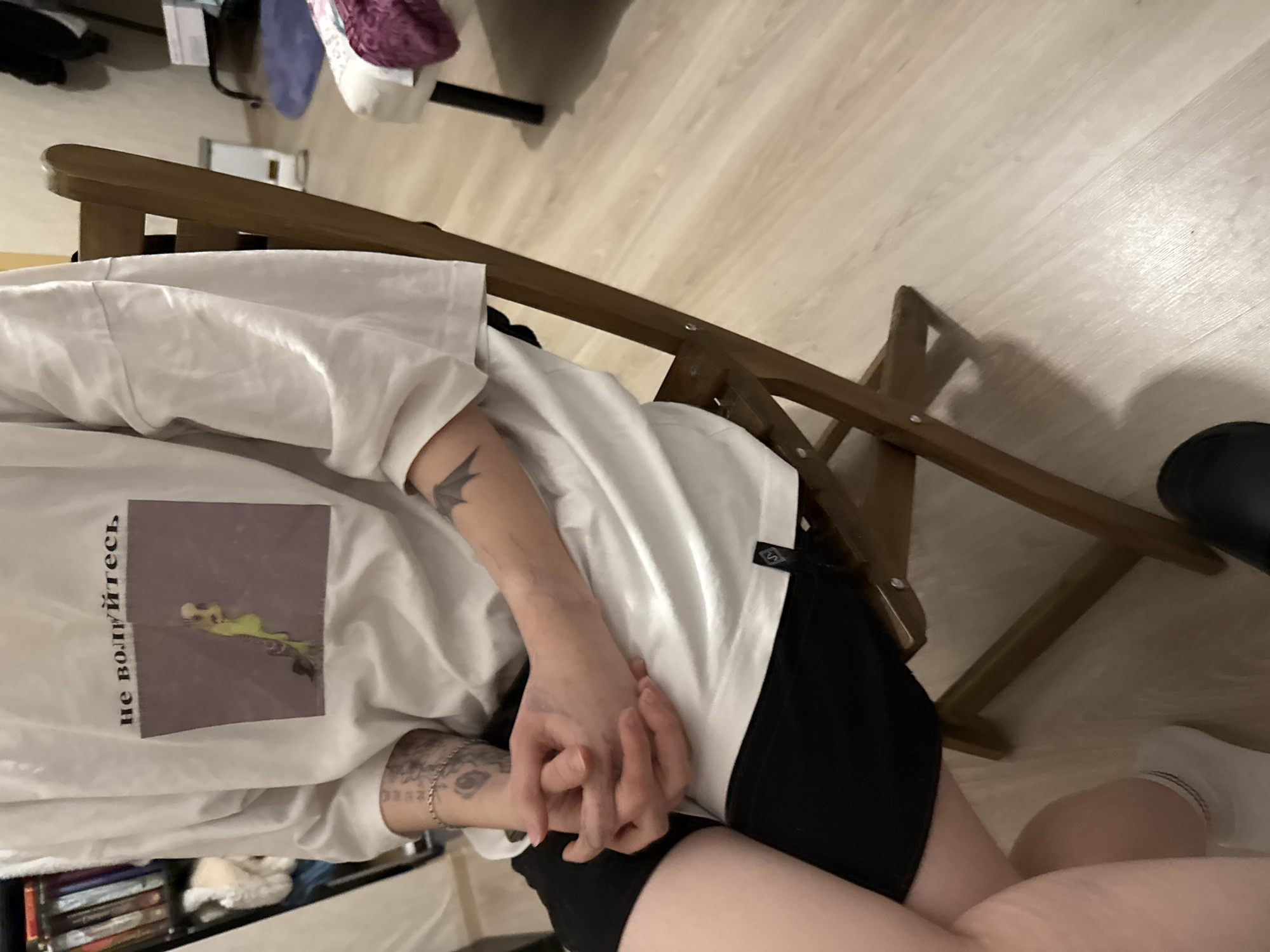 The scene depicts an individual seated in an indoor setting, identified as a room within Saint Petersburg, Russia. The person is partially visible, from mid-thigh up, wearing a white, loose-fitting short-sleeve t-shirt with a graphic on the chest. The graphic displays a rectangular image of a stylized, elongated, greenish-yellow figure on a muted purple background. Above this image, black Cyrillic text reads "не волнуйтесь" (don't worry). A small black label with a white emblem is attached to the t-shirt's lower left hem. The individual also wears black shorts. Tattoos are present on both forearms: the left forearm features multiple designs, including an eye and script, along with a silver-toned chain bracelet; the right forearm displays a bat-like creature tattoo. Both hands are clasped together in the lap. The individual is seated on a light brown wooden folding chair. The flooring is light-toned laminate or wood. In the background, a dark bookshelf filled with books is on the left. To the right, blurred elements include a white rectangular object, a dark metal stand, and an upholstered piece of furniture with a burgundy fabric draped over it. A blue circular or oval rug is also visible on the floor in the background. The visible left foot wears a white ankle sock with dark stripes, while a black slip-on shoe rests on the floor next to the chair.
