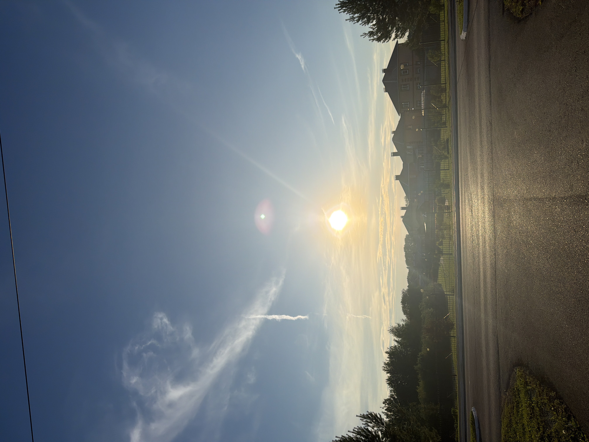 The image depicts a rural or suburban landscape at sunrise or sunset. The sun, a bright orb with a lens flare, is low in the sky, casting a warm, golden light across the scene. Wispy clouds stretch across a clear blue sky, some forming elongated streaks. In the middle ground, a row of houses with dark-tiled roofs are visible behind a black metal fence. Trees, mostly evergreen, line the foreground and the far background, with some shorter trees and bushes interspersed among the houses. A paved road, reflecting the sunlight, occupies the bottom half of the frame, curving slightly on the left side. A thin, dark power line stretches horizontally across the upper portion of the image. The overall impression is one of a peaceful, early morning or late evening in a residential area. There are no people or animals visible in the frame. The location is identified as Shmidovo, Russia.