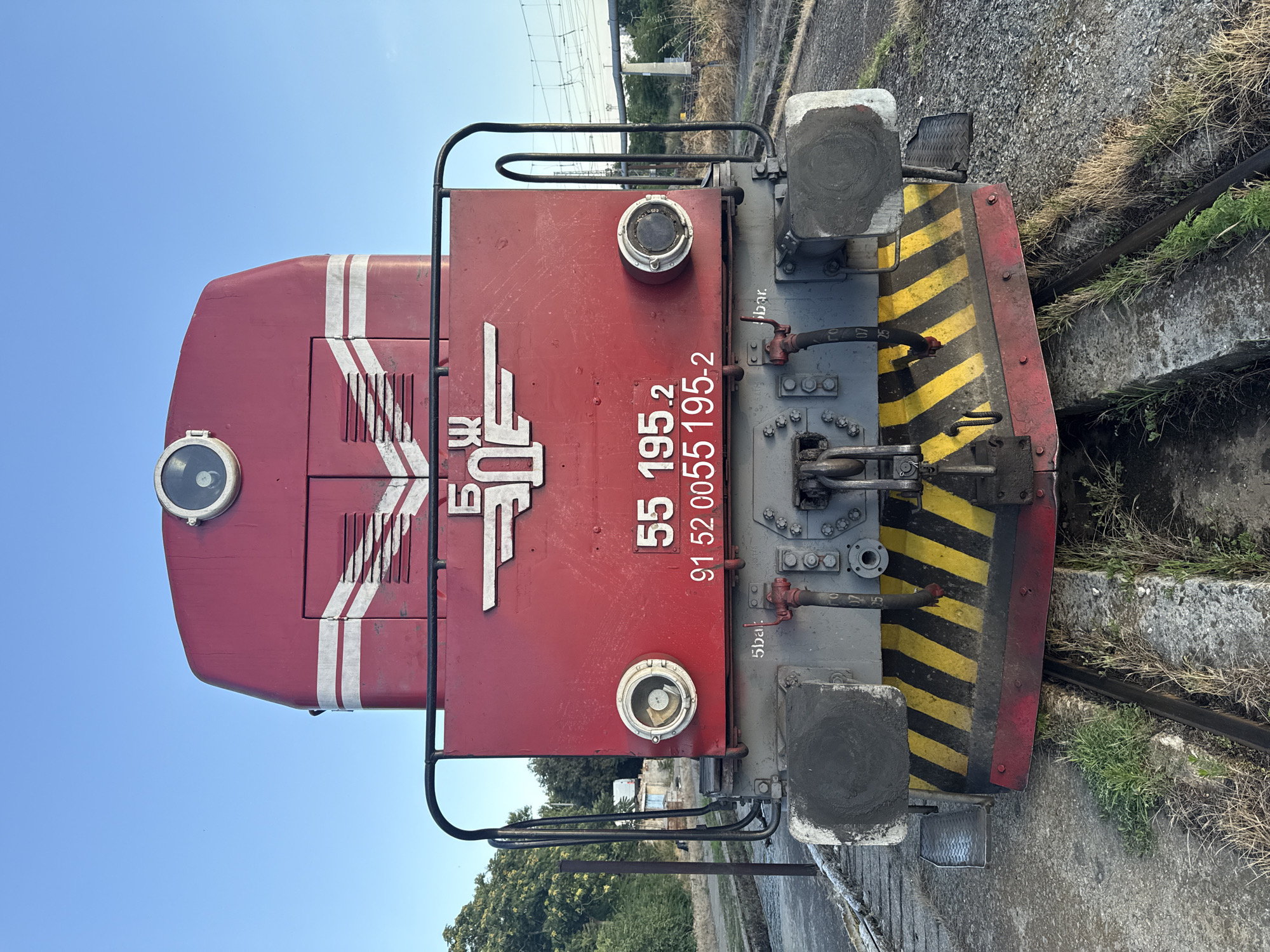 A red diesel locomotive, numbered 55 195.2, is parked on a railway track in Burgas, Bulgaria. The locomotive has a large round headlight at the top, and two smaller round lights on either side of the front panel. The front of the locomotive is marked with white and yellow diagonal stripes, indicating caution. The Bulgarian Railways (BDZ) logo is visible on the front panel, along with the locomotive's identification number. The surrounding environment appears to be a railway yard, with tracks, gravel, and some vegetation visible. The sky is clear and blue.