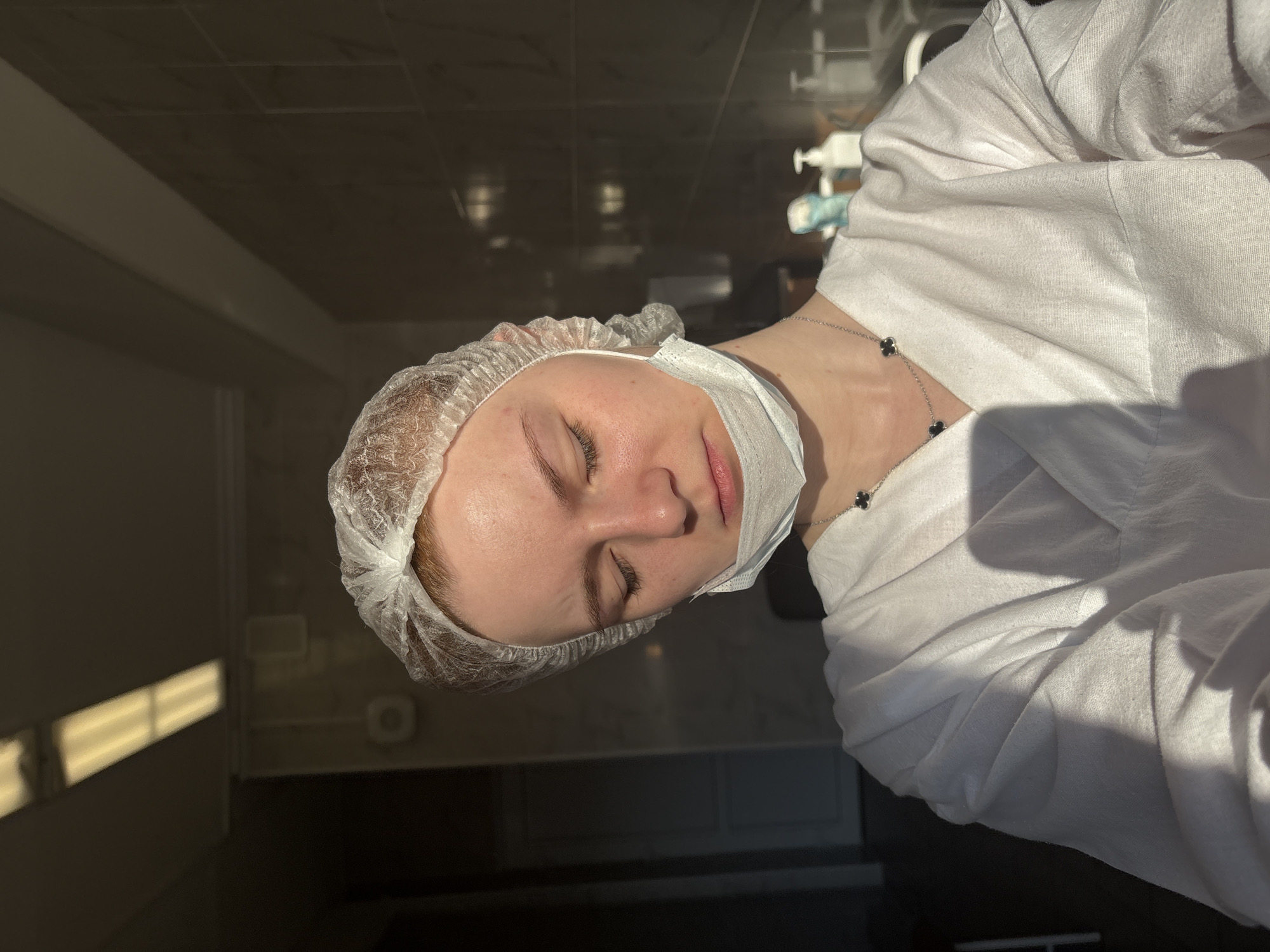 A young person is depicted indoors, likely in a clinic or salon setting in an unknown location in Russia. The individual, who appears to have fair skin and light-colored hair, is wearing a white disposable bouffant cap and a white disposable medical mask, which is pulled down to rest below their chin. They are also wearing a white long-sleeved garment, resembling a lab coat or a treatment gown. A silver necklace adorned with two black clover-shaped charms is visible around their neck. The person's eyes are closed, and their expression appears serene, suggesting they might be resting or undergoing a procedure.
The lighting is bright and directional, casting distinct shadows and reflections on the dark walls and ceiling, indicative of natural sunlight entering the room, possibly during the morning or late afternoon. In the background, partially obscured, is what appears to be medical or cosmetic equipment, including a pump bottle dispenser, reinforcing the professional care setting. The overall atmosphere is calm and clinical. There is no visible text in the image.