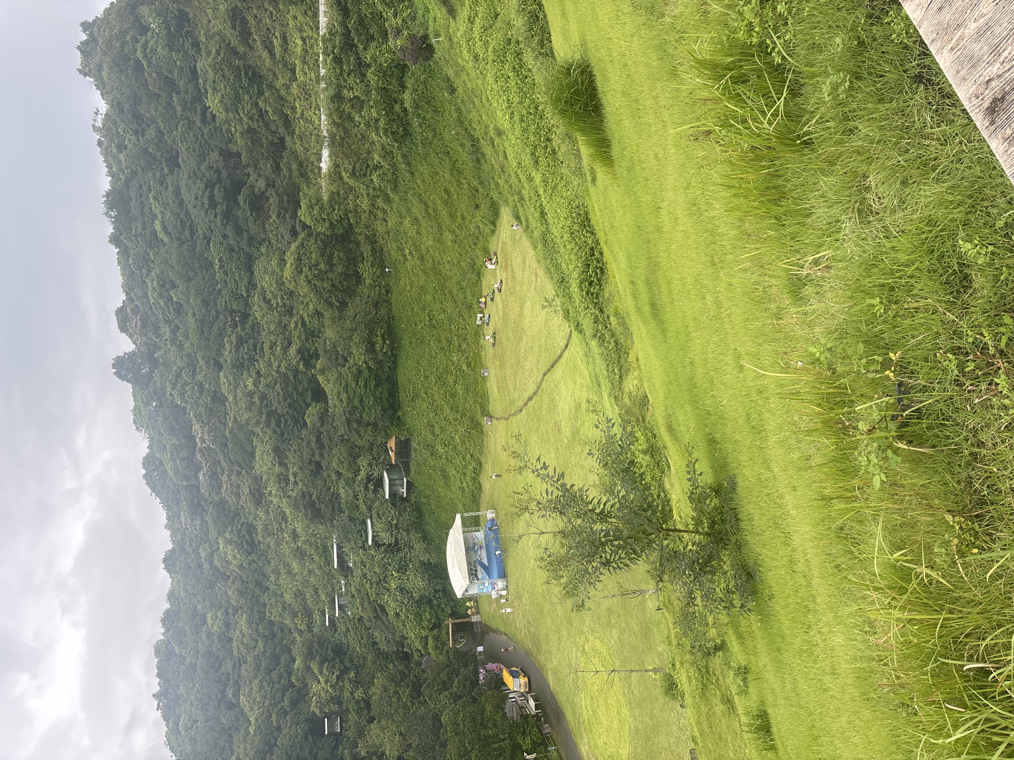 This image captures an outdoor event or gathering taking place in a lush, grassy valley or amphitheater. In the mid-ground, a white-canopied stage with blue accents is set up on a flat grassy area. Several people are visible on and around the stage, and a small yellow vehicle is parked to the left of it. Further into the valley, a group of people are seated on blankets or mats on the grass, suggesting a picnic or an outdoor performance. The surrounding environment is characterized by steep, tree-covered hillsides, indicating a natural or park-like setting. The sky is overcast. The scene appears to be from Guangzhou City, China, given the context provided.