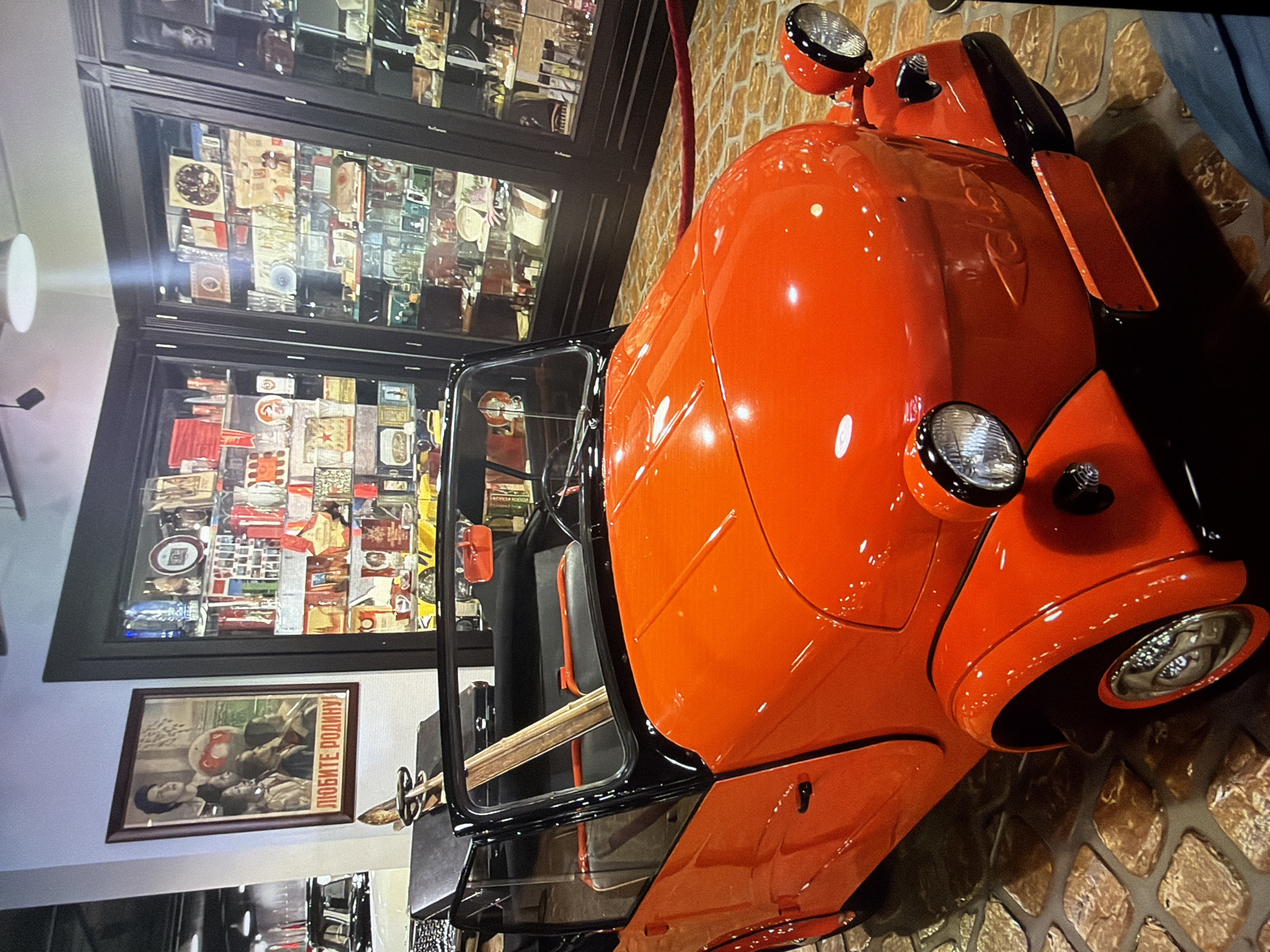 An orange, convertible, two-seater vehicle is displayed on a cobblestone floor inside a museum. The car has a black convertible top, black trim, and round headlights. A wooden plank leans against the windshield. Behind the car, to the left, is a framed poster with Russian text that reads "LUBLIE RODINU!" (Love the Motherland!). To the right of the poster, and behind the car, is a large glass display cabinet filled with various artifacts. The background suggests an indoor exhibition space. The location is identified as the Museum of History and Urban Development in Kotelniki, Russia. There are no people visible in the immediate vicinity of the car.
