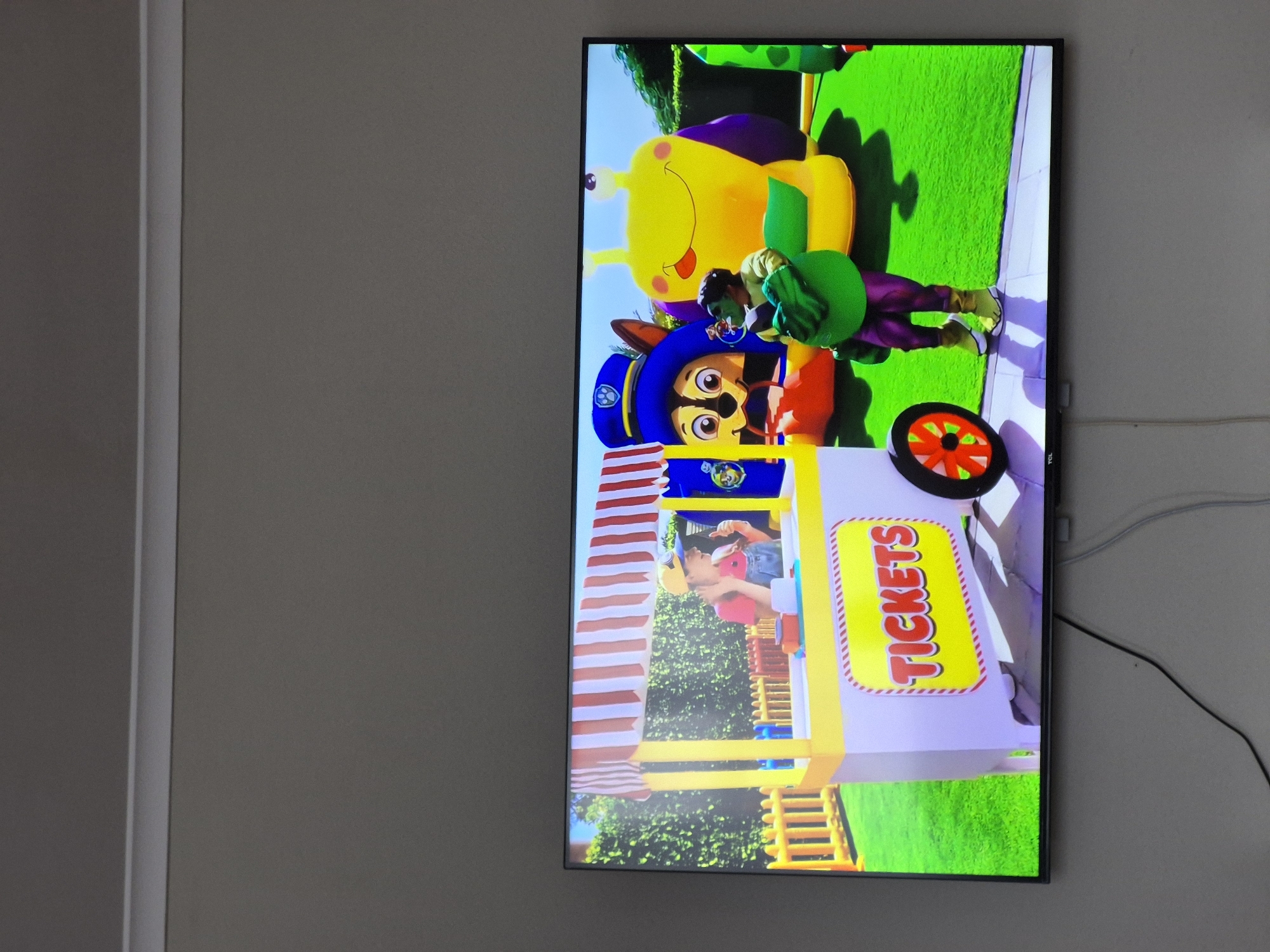 A television mounted on a gray wall displays a cartoon scene featuring a Paw Patrol character, Chase, standing near a ticket booth. Behind Chase, a person dressed in green is interacting with a yellow and purple caterpillar character. The location is not discernible from the image.