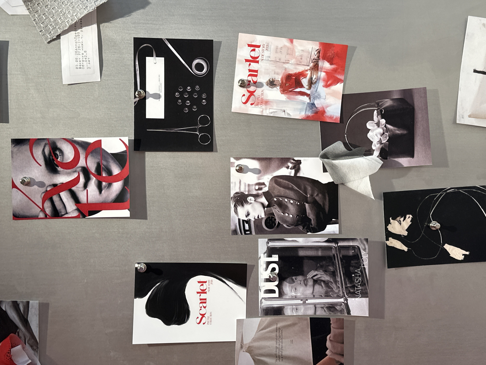 The image displays a mood board or collection of fashion-related inspiration items pinned to a grey, textured wall. The items include magazine covers, photographs, fabric swatches, and handwritten notes. The central elements are two "Scarlet" magazine covers, one featuring a stylized black silhouette and the other a watercolor-style illustration of a woman in red. Other images include a close-up portrait of a woman with red lettering across her face, a black and white photograph of a woman in a military-style jacket, a diptych image of a woman applying makeup, and a photograph of a black handbag filled with white flowers. There is also a collection of tailoring tools, buttons, and a fabric sample. No specific location in Moscow, Russia is identifiable.