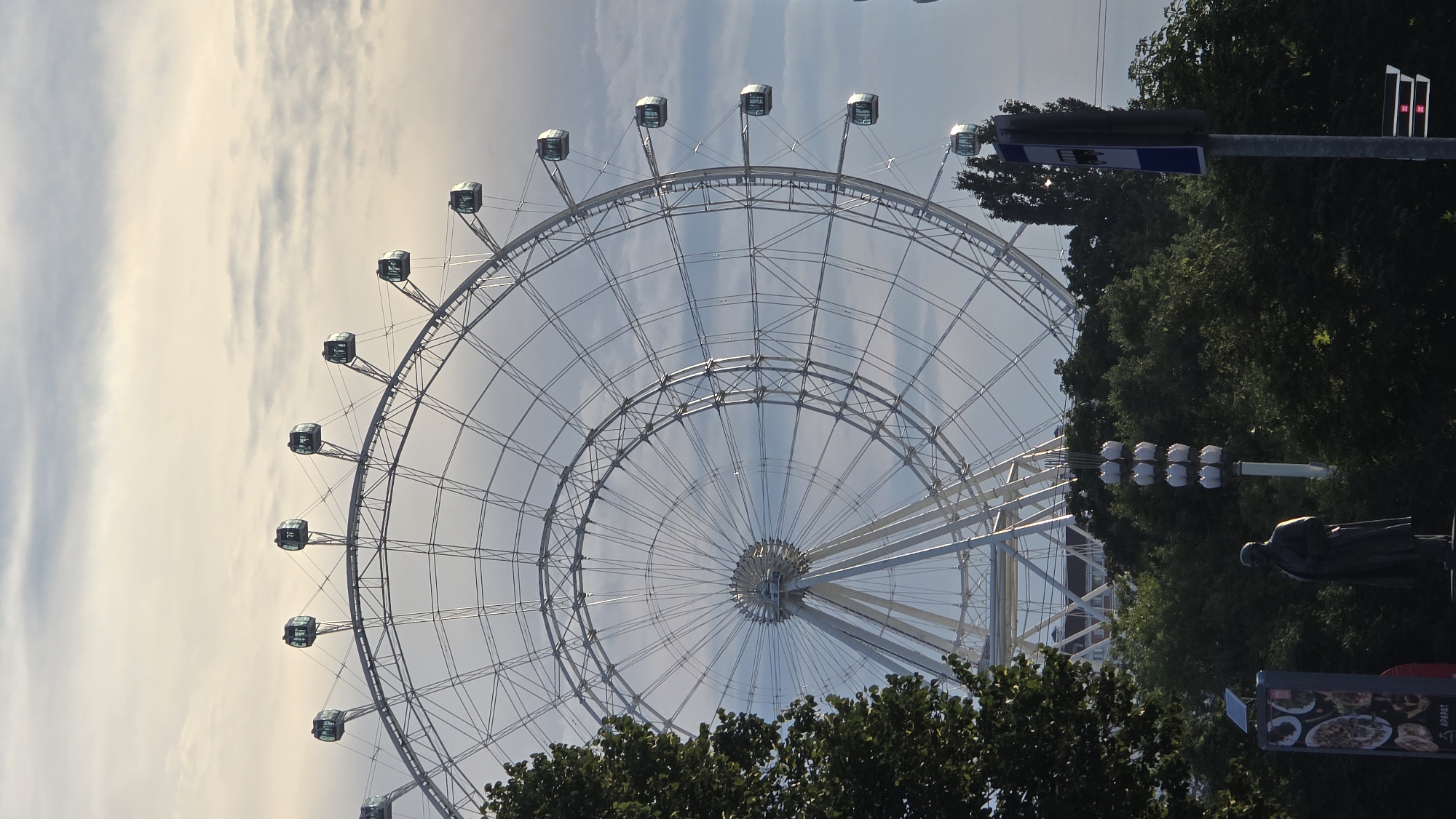 The image depicts a large Ferris wheel in Moscow, Russia, likely at an amusement park or public recreational area. The Ferris wheel is a dominant feature, with its intricate metal structure and multiple gondolas visible against a cloudy sky. In the foreground, dense green trees partially obscure the base of the Ferris wheel. To the left, a statue of a man in a coat stands on a pedestal, and a sign with some text and images is visible next to it. To the right, a bus stop sign is mounted on a pole. The overall scene suggests an outdoor setting, possibly during daytime or early evening, given the soft lighting and cloud cover.