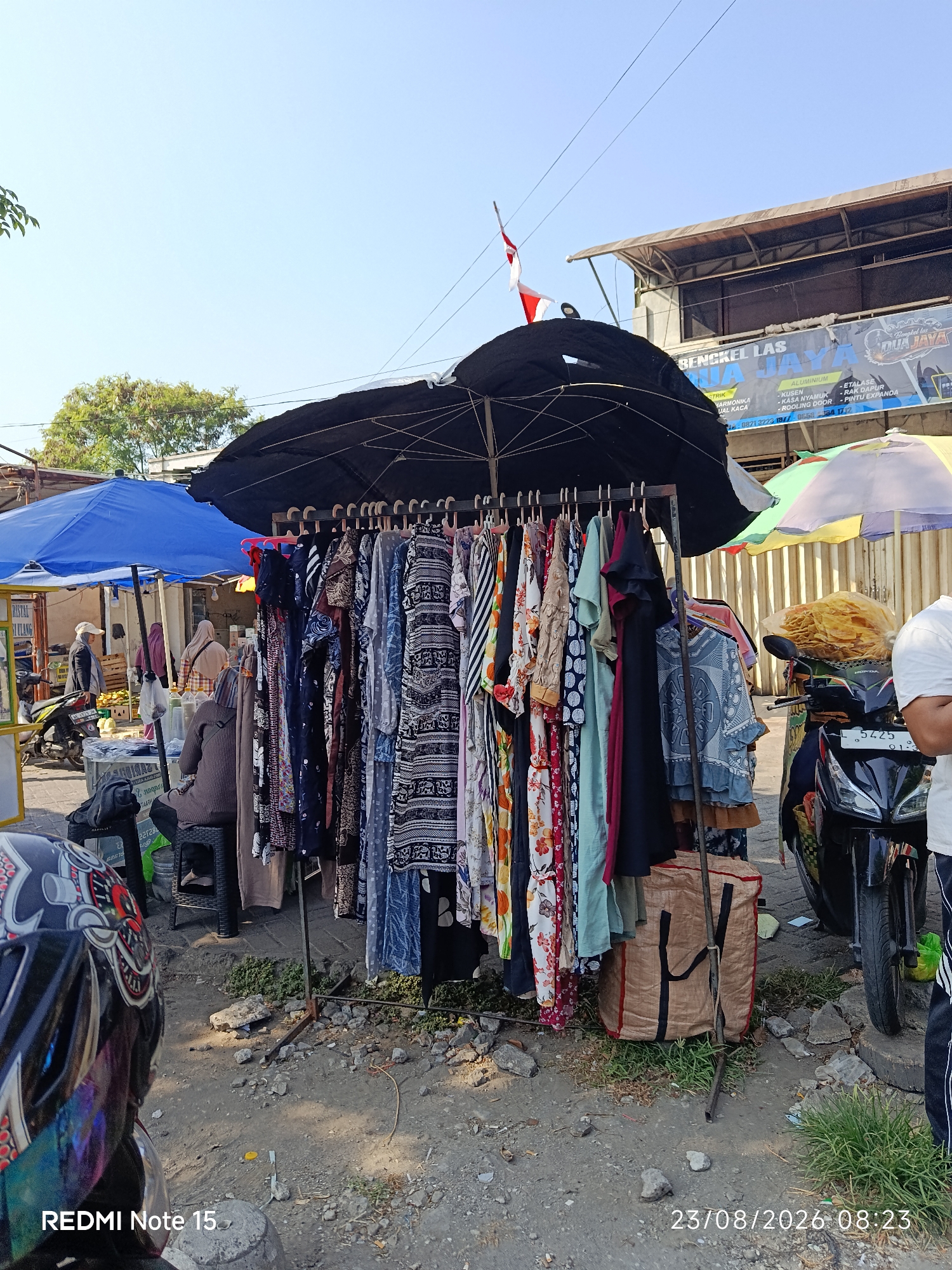 In Keboansikep, Indonesia, an outdoor market stall is displayed under a large black umbrella. Clothing, primarily dresses and shirts, hangs on a rack in front of the umbrella. Some of the clothing features floral and patterned designs. Behind the clothing rack, a brown duffel bag sits on the ground. To the right, a black motorcycle is parked. In the background, a building with a sign that reads "BENGKEL LAS DUA JAYA" is visible. Several people are present in the scene, including a person in the foreground wearing a motorcycle helmet and another person on the right side of the image wearing a white t-shirt and dark pants. The ground is a mix of dirt and gravel, with some patches of grass. The sky above is clear and blue. A date and time stamp in the lower right corner reads "23/08/2026 08:23".