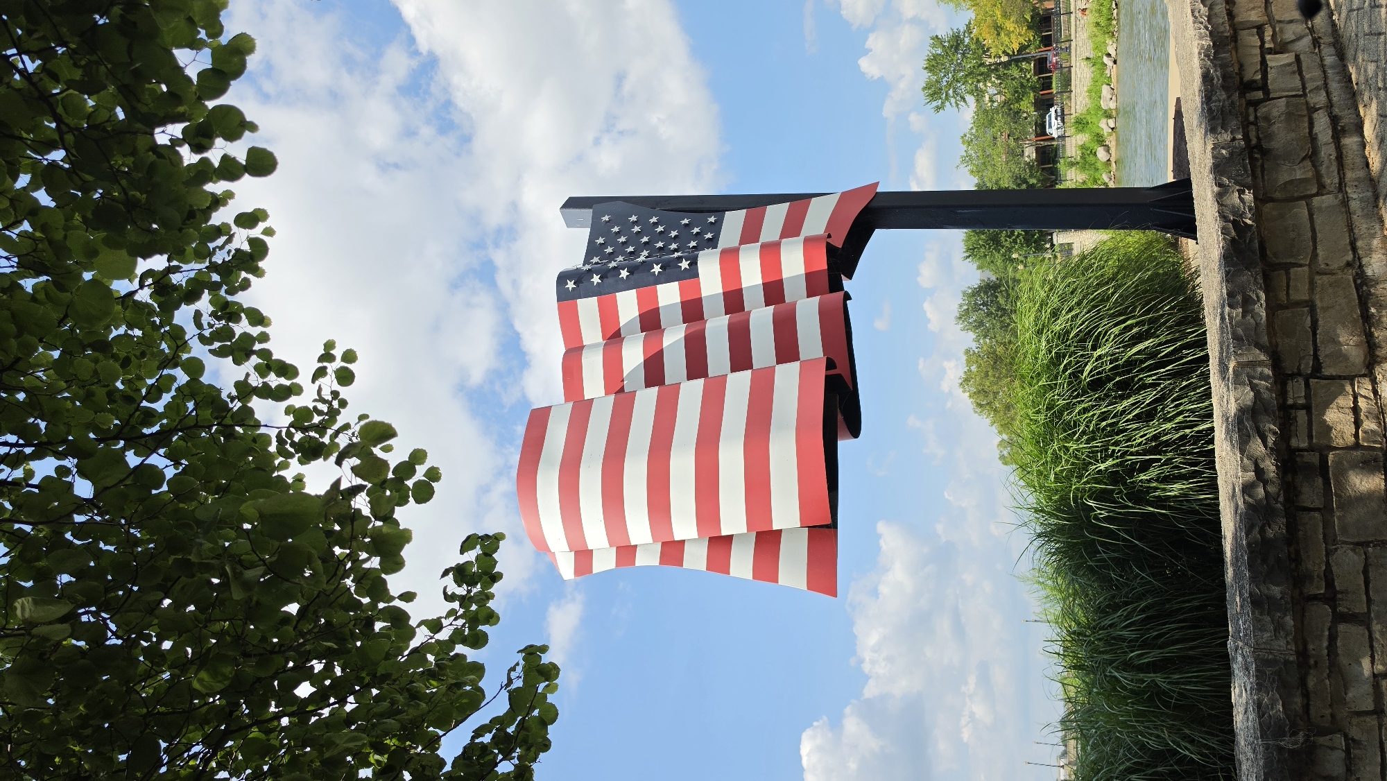 In Elgin, United States of America, a large, stylized sculpture of the American flag stands tall against a bright blue sky dotted with fluffy white clouds. The flag, rendered in bold red and white stripes and a blue union with white stars, appears to be waving dynamically, its fabric-like sections folded and curved. The sculpture is mounted on a dark, sturdy pole, which is anchored to the ground behind a low stone wall. In front of the wall, lush green ornamental grasses create a verdant border. Beyond the grasses and the flag, a body of water, likely a river, is visible, with rocky banks and trees lining the opposite side. In the background, a few buildings and trees suggest an urban or suburban setting. The image is captured from a low angle, with the branches of a leafy green tree partially framing the top of the scene.