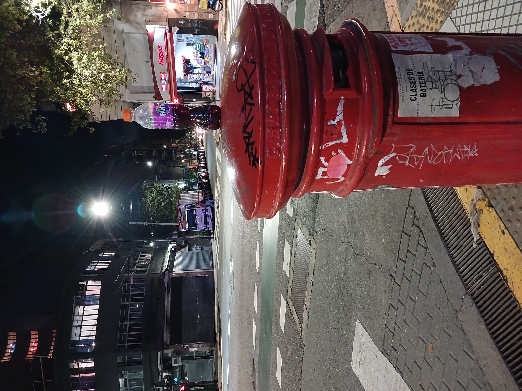 A nighttime street photograph, oriented vertically, captures a vibrant red, traditional-style mailbox in an urban setting, likely Buenos Aires, Argentina. A purple Fanta bottle rests prominently on top of the mailbox.
The mailbox is adorned with various graffiti tags in black and white, including partially visible Arabic words like "عاجل" (urgent) and "حميد" (Hamid). A white poster is attached to its side, advertising "CLASES DE BATERÍA" (Drums Classes) by "FRANCO," complete with a phone number "1138465199" and additional text indicating "TODAS LAS CUADRAS..." and "ZONA CERTOO."
The scene is illuminated by artificial street lighting, with a bright streetlight in the mid-ground creating a noticeable lens flare. In the background, buildings line the street, one of which features a brightly lit storefront with an "Energizer" sign visible. Trees with illuminated foliage are also present. The street itself has clear white and green crosswalk markings, and the sidewalk features textured tiles and a metal drainage grate. The atmosphere suggests a quiet urban evening.