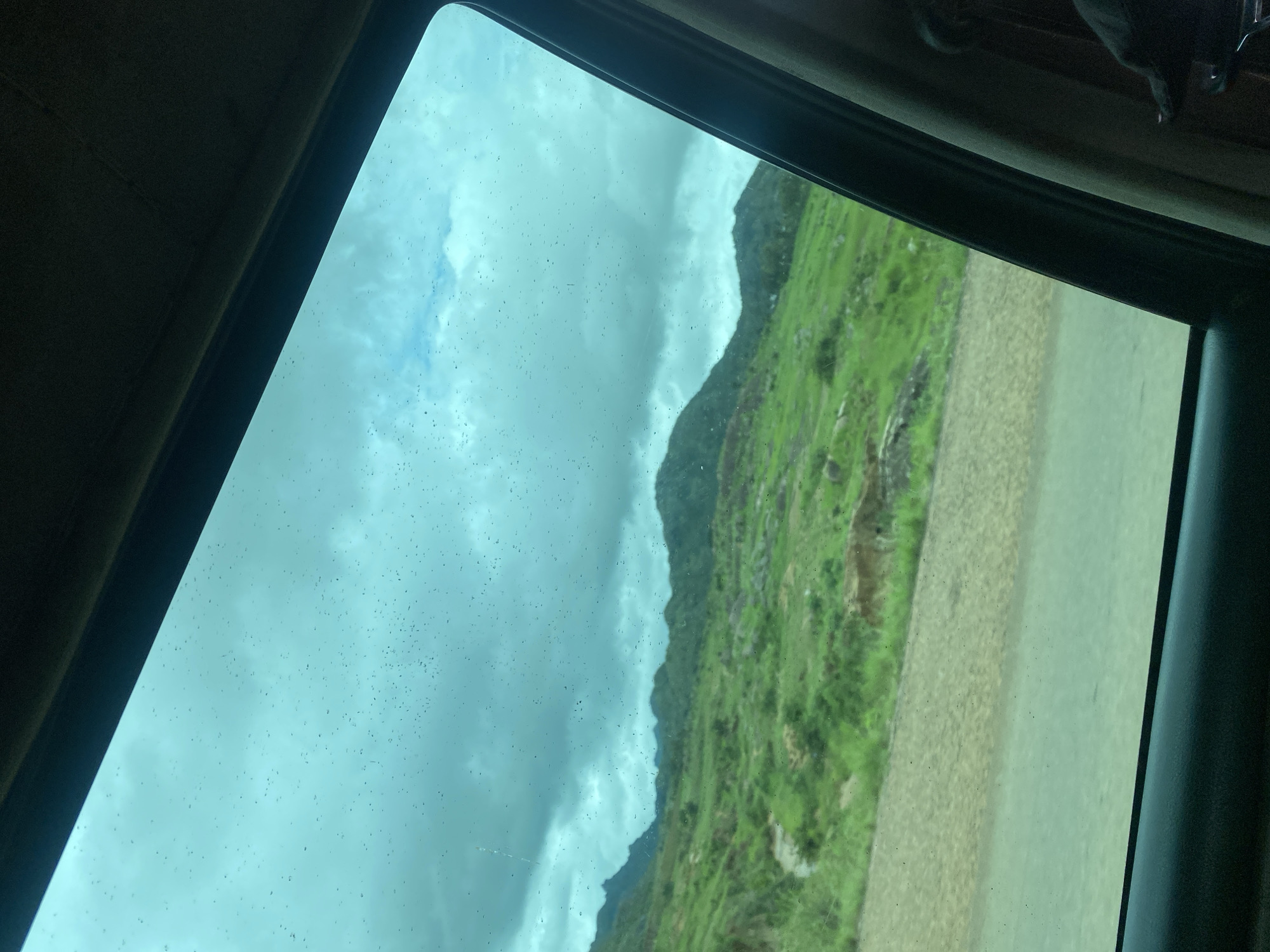 The image, taken from inside a vehicle, shows a landscape viewed through a tinted window. The window is speckled with small dark spots, possibly dirt or water droplets. The scene outside consists of rolling green hills with patches of brown earth and rocks. In the distance, darker, more rugged mountains are visible under a cloudy, teal-colored sky. A paved road runs horizontally across the lower portion of the frame, with a gravelly area bordering it on the left. The interior of the car, including a dark fabric-covered A-pillar and a portion of a seat, is visible on the right side. There is no indication of the specific location within Wang, Nigeria.