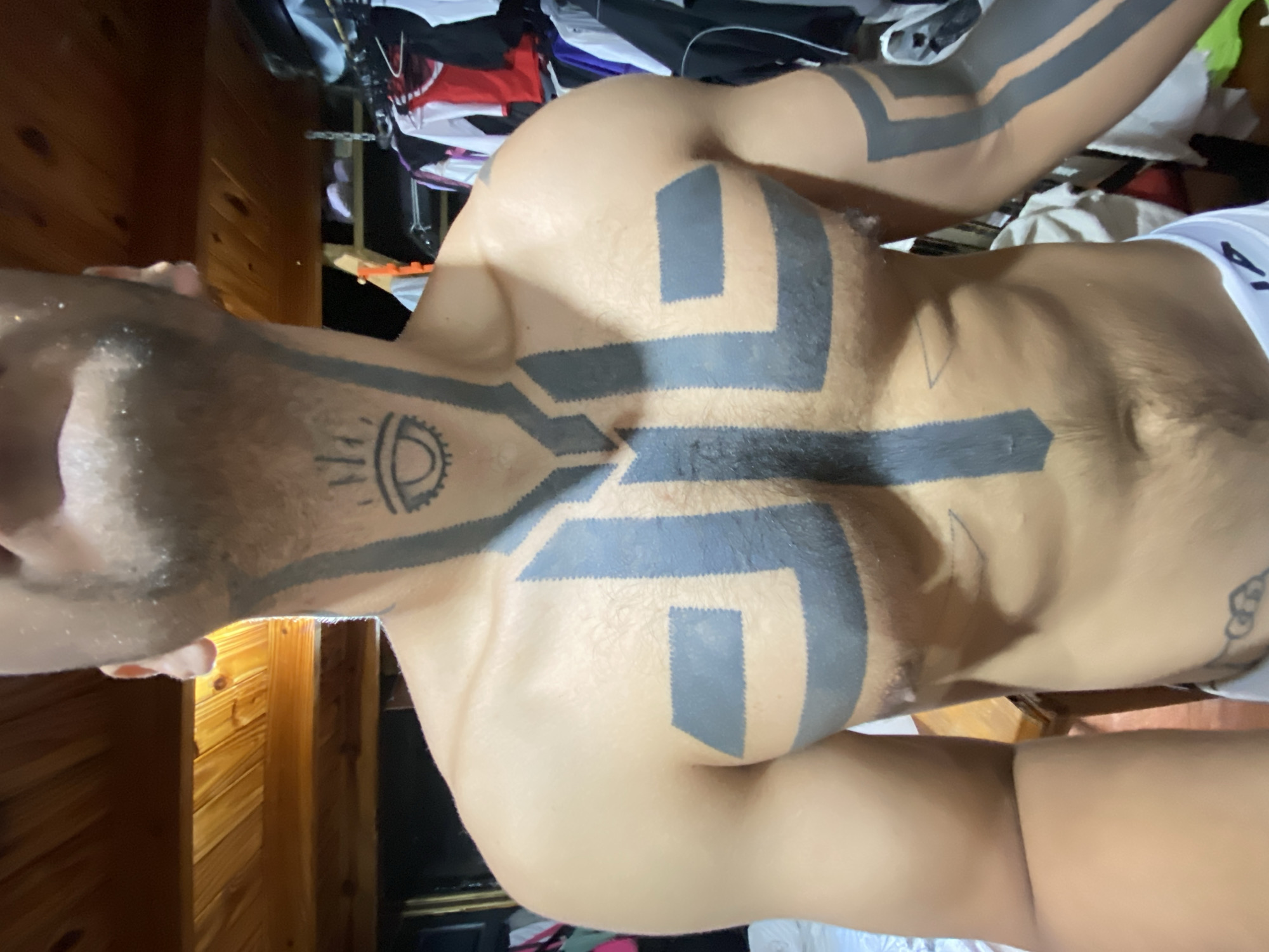 A close-up, angled view depicts a person's torso and upper arms, heavily tattooed with thick, dark geometric designs. The tattoos extend across the chest, shoulders, and down the arms. The person has a light complexion and some visible body hair. Their face is partially visible, showing a damp or sweaty complexion. The background includes wooden paneling on a wall and a cluttered shelf with clothing items. The exact location in Buenos Aires, Argentina is not discernible from the image provided.