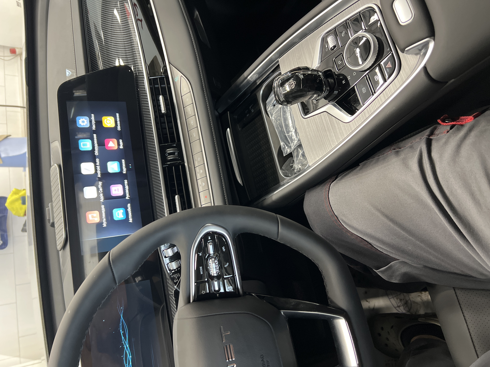The image depicts the interior of a modern vehicle, specifically from the driver's perspective, located in Izhevsk, Russia. The scene reveals a luxurious and technologically advanced cabin.
In the foreground, the upper portion of a person's left thigh, clothed in grey trousers with a red stitch detail, is visible. Below the steering wheel, the person's left foot, wearing a grey Crocs-style shoe, rests near the accelerator pedal.
The multi-function steering wheel, upholstered in black leather, occupies the left side of the frame. The left spoke features various controls, including a "MODE" button, a rotary dial, and buttons for phone calls and other functions. The text "JET" is visible on the airbag cover.
Dominating the dashboard is a large, integrated digital display system. The screen on the left, partially obscured by the steering wheel, shows a blue graphical interface. The central infotainment screen displays a grid of application icons with Russian labels: "Мультимедиа" (Multimedia), "Apple CarPlay," "Android Auto," "Оборудование" (Equipment), "Настройки" (Settings), "Автомобиль" (Car), "Руководство" (Manual), "Картинки" (Images), "Видео" (Video), and "Обновление" (Update). The dashboard and door trim feature a carbon fiber pattern.
Below the infotainment screen, a row of climate control buttons is present, including "A/C" and "AUTO" functions. The central console, finished with a brushed metallic texture, houses a distinctive crystal-like gear selector adorned with a stylized emblem. Surrounding the shifter are various control buttons, including "P," "R," "N," "D," an electronic parking brake, a hazard light button, and a rotary controller labeled "MODE." In a storage compartment forward of the gear selector, a clear plastic bag containing car keys (a key fob and a metal key) is visible.
Reflections in the windshield and dashboard reveal an indoor environment with white tiled walls and overhead lighting, along with a yellow cloth and a blue object, suggesting the vehicle is in a garage, service bay, or showroom.