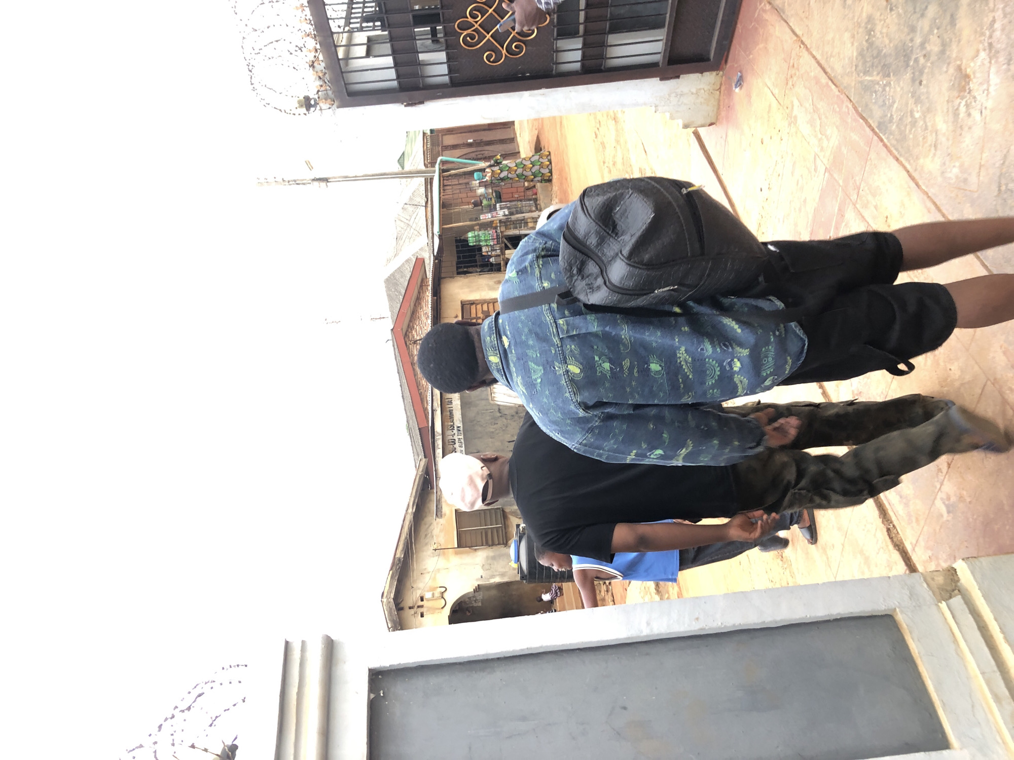 The image, rotated 90 degrees clockwise, captures an outdoor street scene under bright daylight. In the foreground, two men are visible from behind. The man on the left wears a blue patterned long-sleeved shirt, a black backpack, and black shorts. Next to him, another man wears a black t-shirt and a light-colored cap, with dark-colored pants. They are standing on a tiled or paved surface that transitions to a dirt path further into the scene.
To the upper left, a black metal gate with an ornate golden design is visible, topped with barbed wire. In the background, several buildings line the street. One prominent building has legible text painted on its facade: "SUPREME AL-ASLIYYAH SCHOOL JIDDAH EFDE TOWN". Near this building, a person in a colorful patterned dress is partially visible next to what appears to be a small market stall or shop. The overall setting suggests a bustling, possibly urban or semi-urban environment under clear, sunny skies.