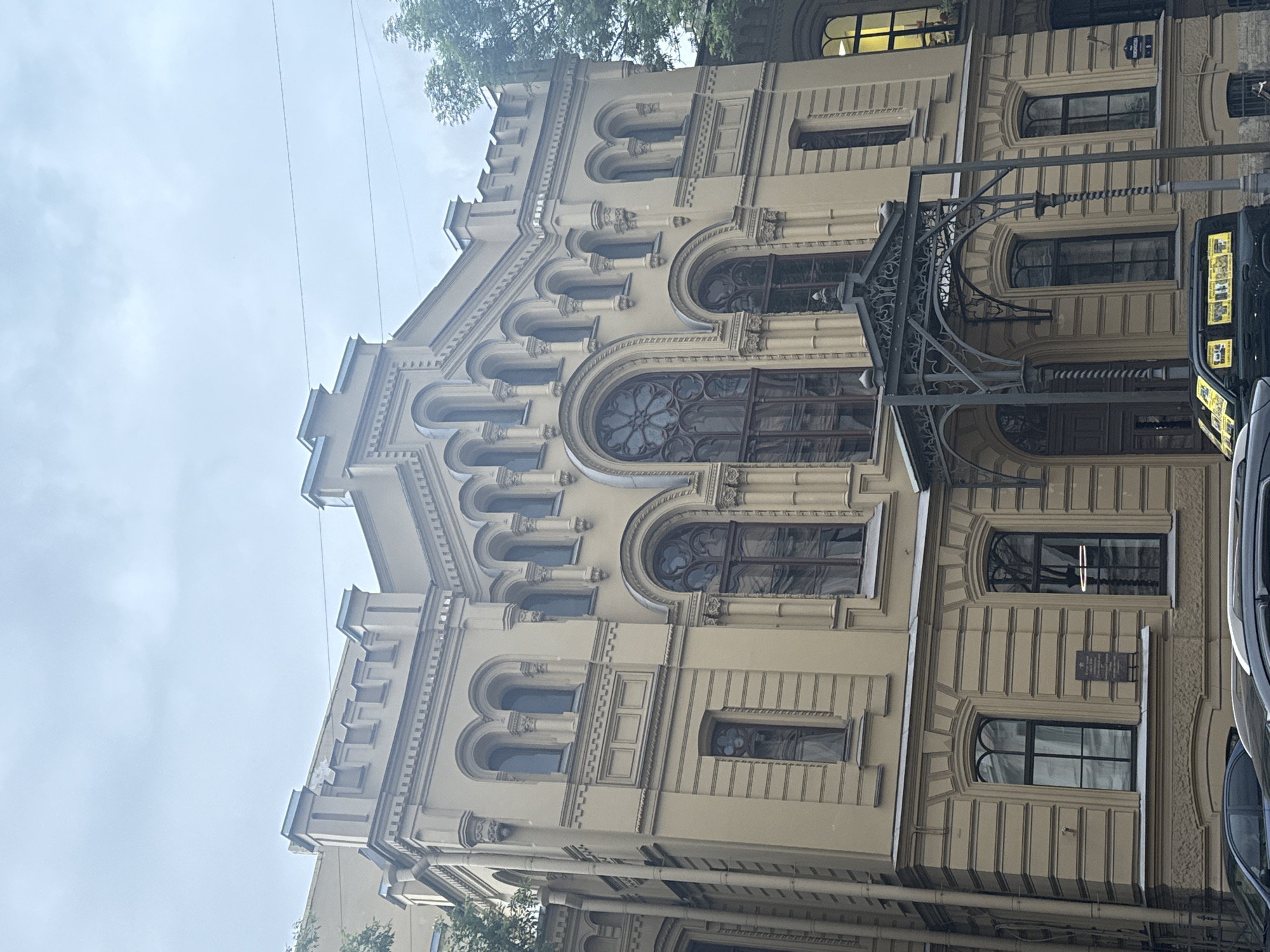 The image shows the exterior of a light yellow, historic building located in Saint Petersburg, Russia. The building has a detailed facade with multiple arched windows, decorative cornices, and a prominent ornate entrance canopy supported by decorative columns. Some windows feature intricate tracery. A dark, modern SUV with yellow posters on its rear window is parked in front of the building, partially obscuring the sidewalk and street. The sky is overcast and cloudy. Trees are visible on the right side of the building. A street sign is partially visible on the right side of the building.