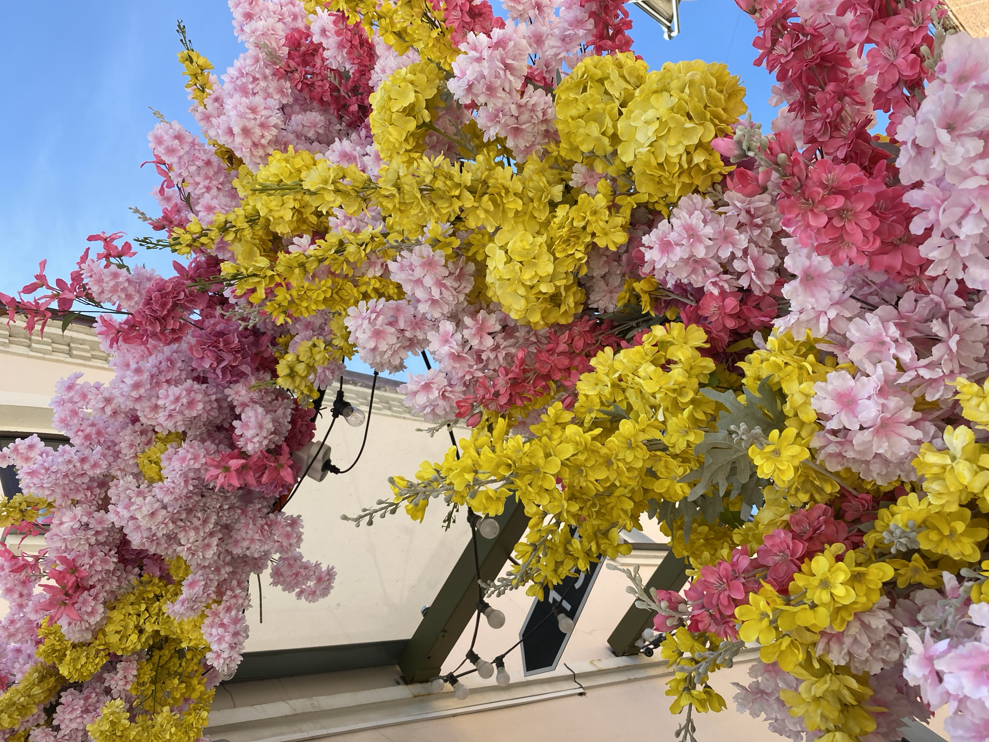 A low-angle, full outdoor shot shows a colorful archway of pink, yellow, and light pink flowers beneath a pale blue sky in Izhevsk, Russia. The floral arrangement is suspended from an awning adorned with strings of exposed light bulbs. A white building is visible behind the awning.