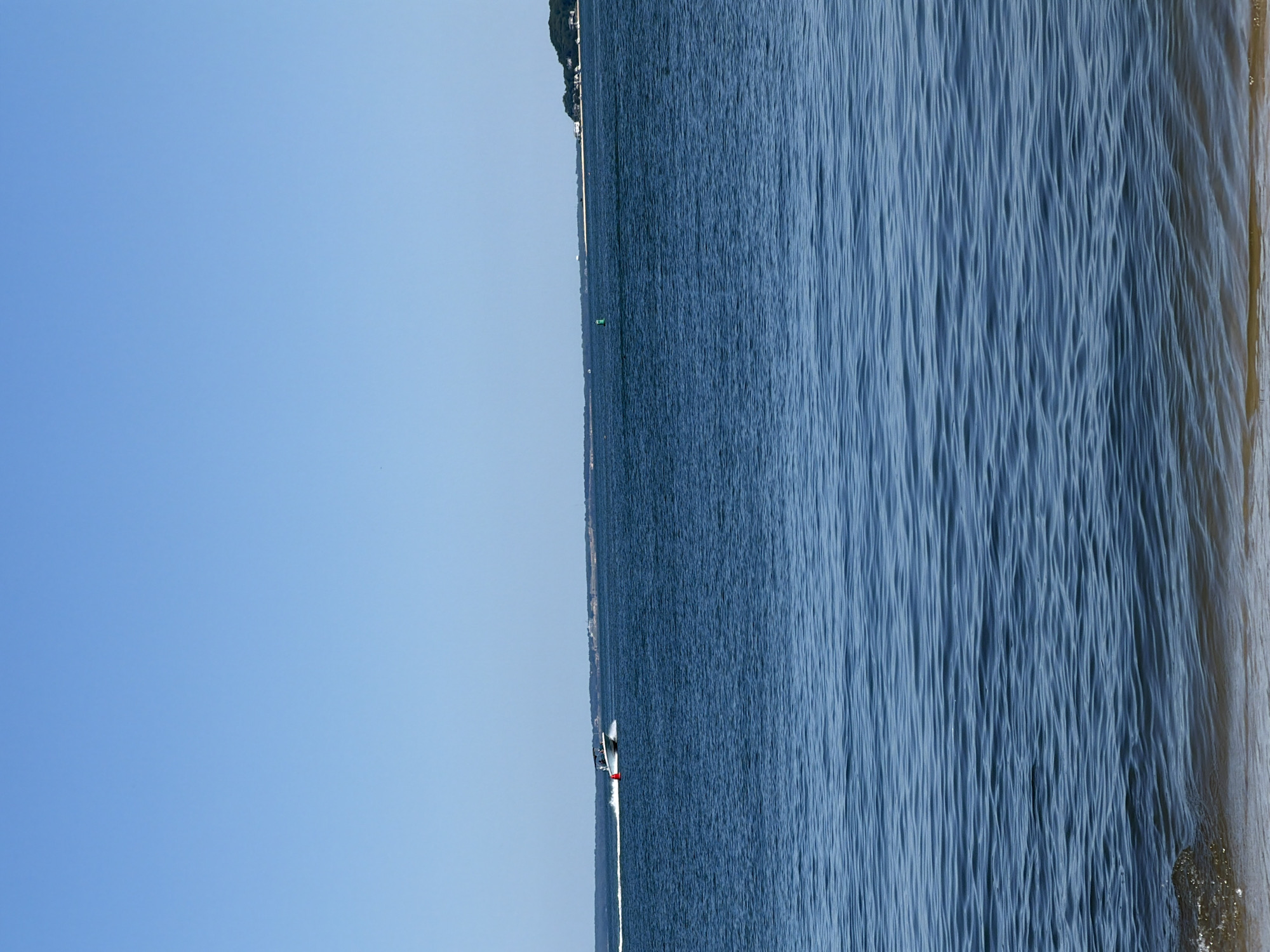 A motorboat speeds through the blue water of a bay under a clear sky. The boat, white with red accents, leaves a white wake behind it. A red buoy floats in the water to the right of the boat. In the distance, a tree-lined shore with scattered houses is visible. The foreground shows the sandy bottom of the bay. The scene appears to be in Riverhead, New York, USA.