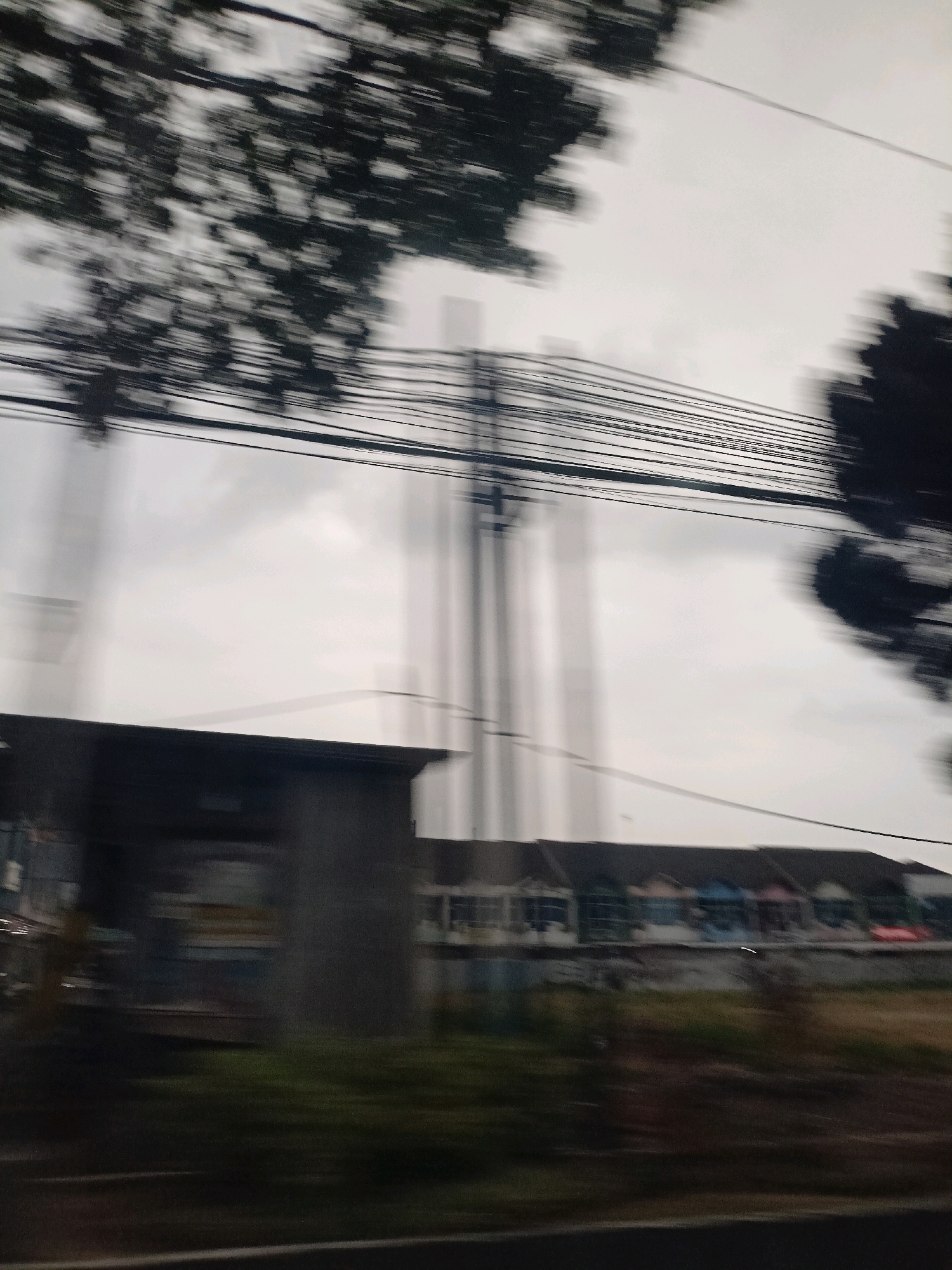 The image depicts a blurred street scene, likely taken from a moving vehicle, in the city of Magelang, Indonesia. In the foreground, a dark, out-of-focus structure is visible on the left. Behind this, a row of colorful houses with dark roofs stretches horizontally across the frame. Overhead, a dense network of power lines runs across the cloudy sky, with the silhouettes of trees visible on the left and right edges of the image. The overall impression is one of motion and a typical urban or suburban environment.