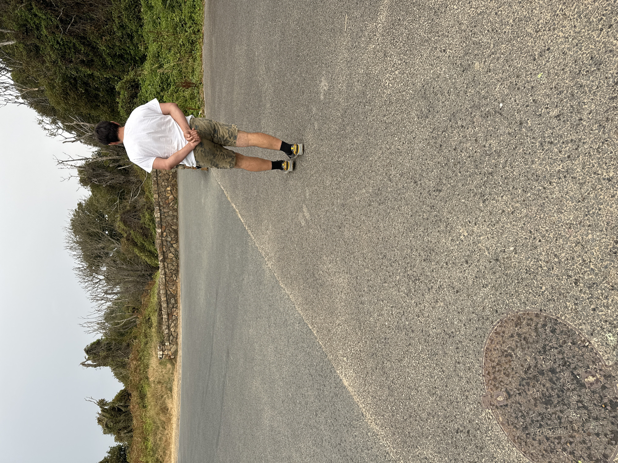 A man wearing a white t-shirt, camouflage shorts, black socks, and athletic shoes stands on an asphalt surface with his back to the camera. He has his hands clasped behind his back. The asphalt is divided by a diagonal line, with one side appearing darker than the other. In the lower left corner of the image, there is a circular, metal manhole cover. Behind the man, a stone wall separates the asphalt from a vegetated hillside with trees and bushes. The sky is overcast. The location is Courtown, Ireland.