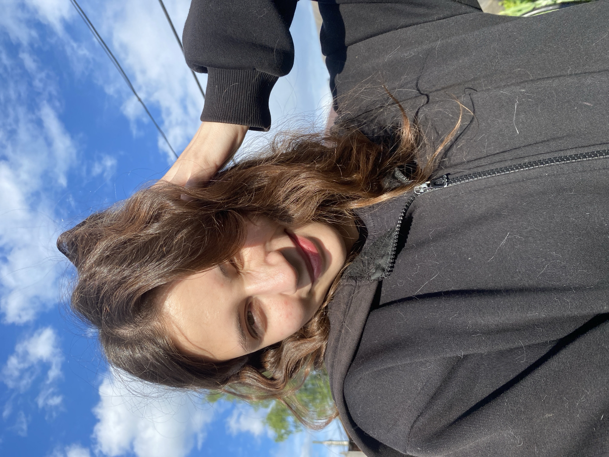 A young woman with brown hair is pictured from a low angle, likely a selfie. She wears a black zip-up hoodie. The sky is visible in the background, with scattered clouds. The location cannot be determined as Votkinsk, Russia, or any other location with certainty. No specific buildings or landmarks provide location clues.