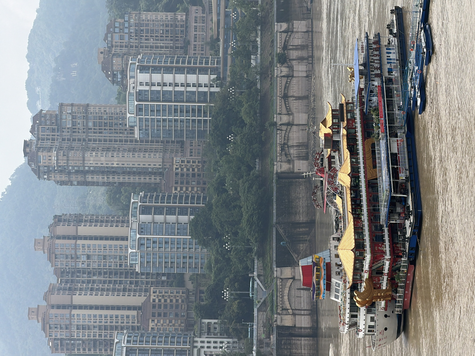 The image depicts a hazy, overcast scene of the Yangtze River in Chongqing, China, likely from Yuzhong District, characterized by a dense urban landscape with numerous high-rise buildings and a distinctive, ornately decorated cruise ship.
In the foreground, a large, multi-tiered cruise ship with traditional Chinese architectural elements dominates the scene. The ship features multiple decks with red and gold detailing, elaborate tiled roofs in yellow, and decorative golden sculptures. Smaller, blue boats are docked alongside or in front of the main vessel. The water is a murky brown, indicative of a river.
Behind the ship, a textured stone embankment or wall lines the riverbank, with arched structures suggesting an older architectural style. Further back, lush green trees form a dense line, separating the riverfront from the cityscape. Scattered among the trees are lampposts with multiple lights.
The background is filled with a dense collection of modern high-rise apartment buildings and towers, varying in architectural style and color, from light beige to reflective glass facades. Some buildings have a stepped or terraced design. In the far distance, hazy, tree-covered mountains rise, their details softened by the atmospheric conditions. The overall impression is one of a densely populated urban environment situated along a major river, with a blend of traditional and modern elements.