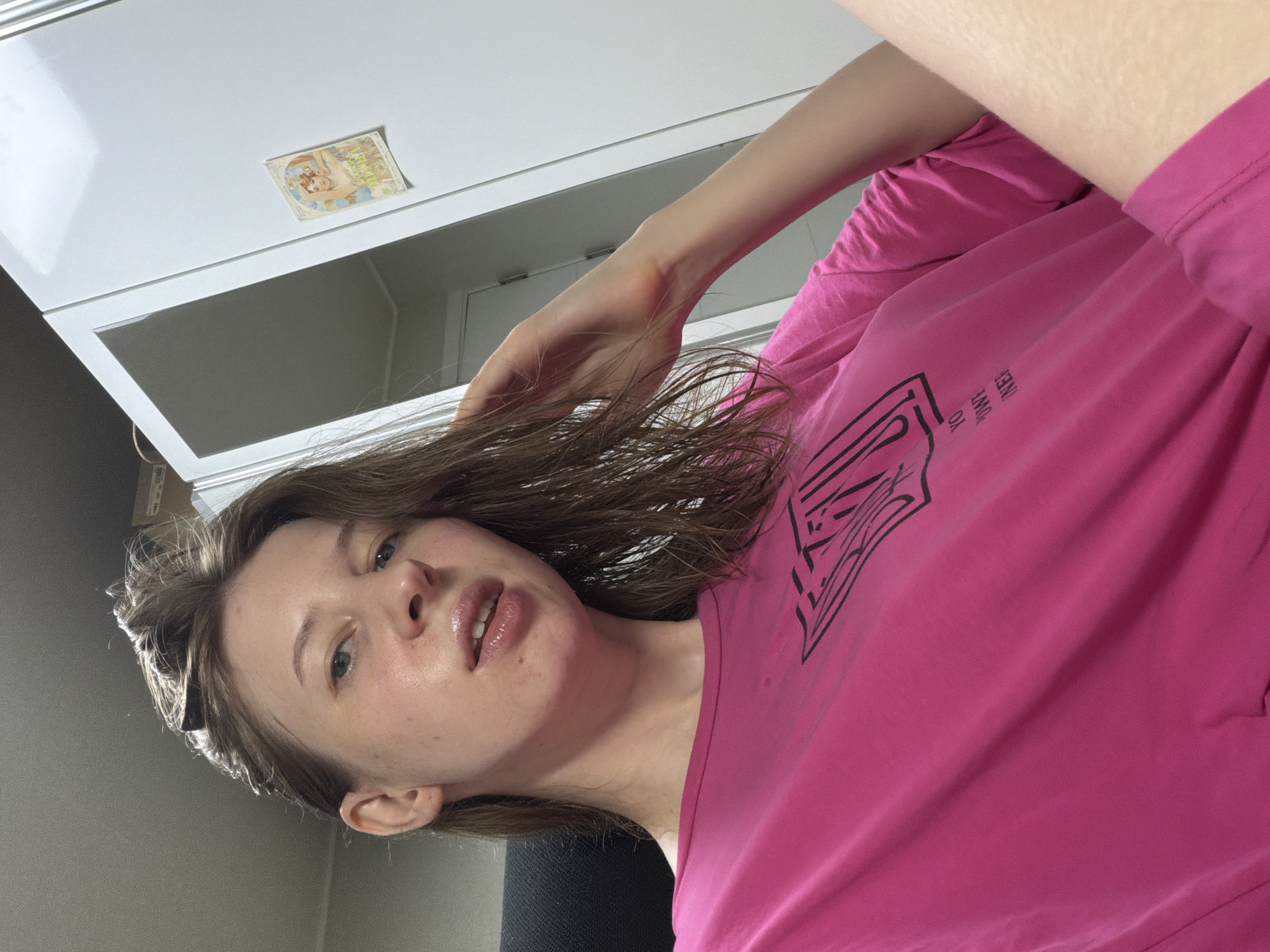 A young woman with light brown hair, wearing a bright pink t-shirt with a graphic design, is seated in an indoor setting. Her hair appears damp and is styled loosely. She is looking towards the camera with a slight smile and her lips are slightly parted. Her right hand is raised, touching her hair near her temple. The background shows a white cabinet or closet door with a small decorative sticker featuring a woman. The lighting suggests an interior environment, possibly a room in a residence. The image does not provide explicit clues to pinpoint its location within Dongducheon-si, South Korea.