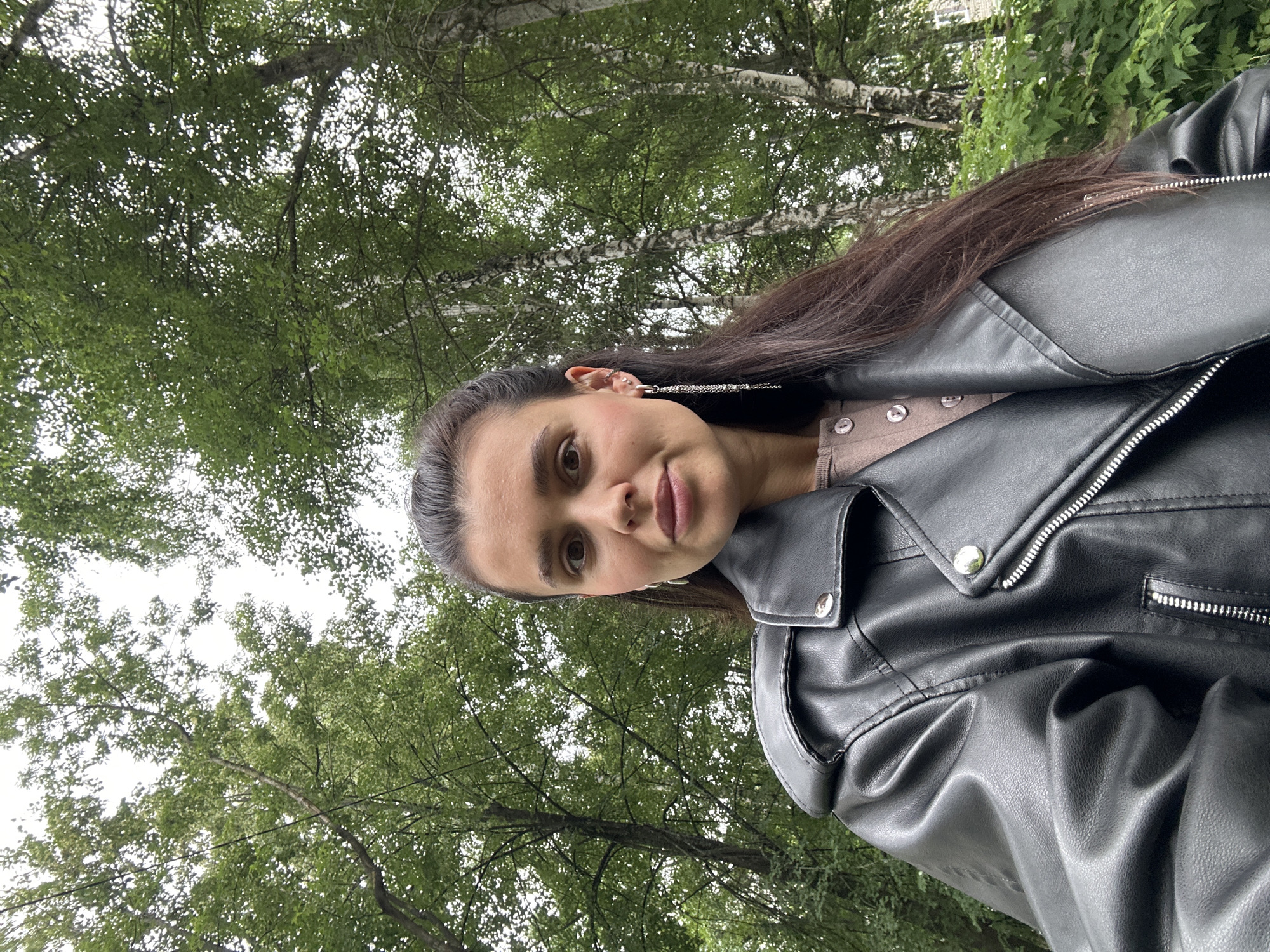 The media file is a portrait of a young woman taken outdoors, likely in Chaikovsky, Russia. She is wearing a black leather jacket over a light brown shirt and has long, dark hair. Her face is partially visible, and she appears to be looking at the camera with a neutral expression. She is wearing a long, silver earring in her visible ear. The background consists of lush green trees with dappled sunlight filtering through the leaves, suggesting an overcast or late afternoon setting. The image is oriented vertically, with the woman's head positioned towards the left side of the frame. There is no visible text in the image. The overall impression is a casual outdoor portrait.
