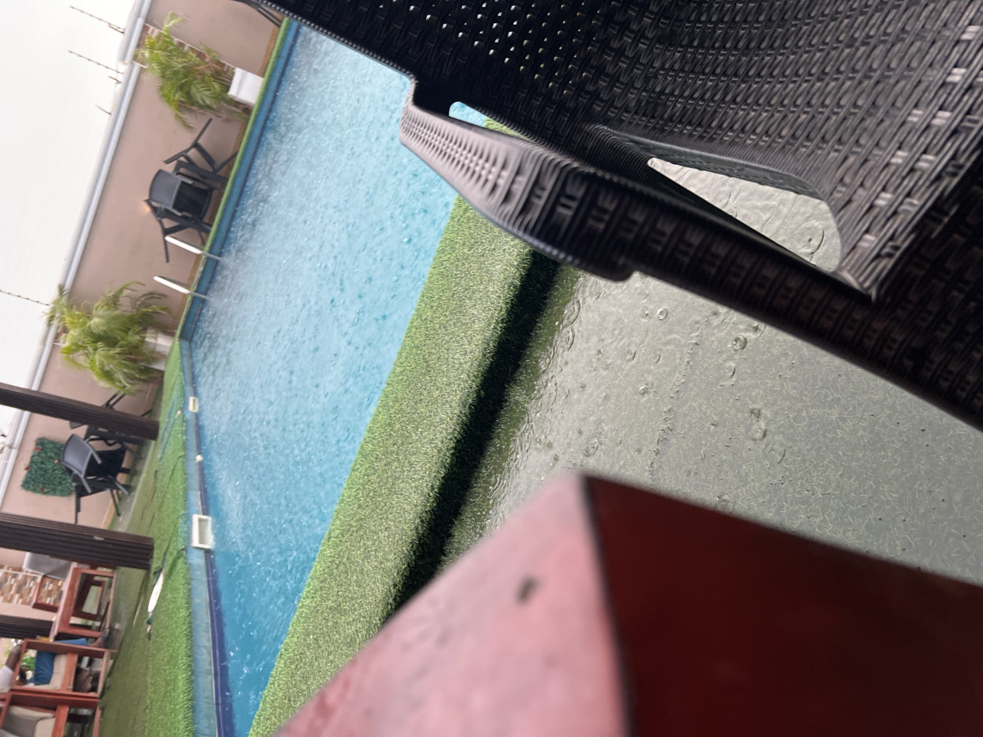 The image captures an outdoor poolside scene in Abuja, Nigeria, viewed from a slightly elevated and tilted perspective. The primary focus is a swimming pool with clear blue water, upon which numerous raindrops are visibly falling, creating ripples and splashes, indicating ongoing precipitation.
In the foreground, part of a dark, woven wicker-style chair is prominent in the upper right. The ground directly in front of this chair and around the pool appears wet, with individual raindrops clearly visible on its surface. A blurred red object occupies the bottom right corner of the frame.
The pool is bordered by a vibrant strip of artificial green grass. Beyond the pool, against a light brown wall, are several potted palm plants and two black, stackable pool chairs. To the left of the pool area, a section with wooden tables and chairs, likely part of an outdoor dining or lounge area, is partially visible under a slatted overhead structure.
The lighting suggests an overcast daytime, typical for a rainy period. The scene is devoid of clearly visible people. The image provides a tranquil, atmospheric snapshot of a poolside area experiencing rainfall. No discernible text is visible within the frame.