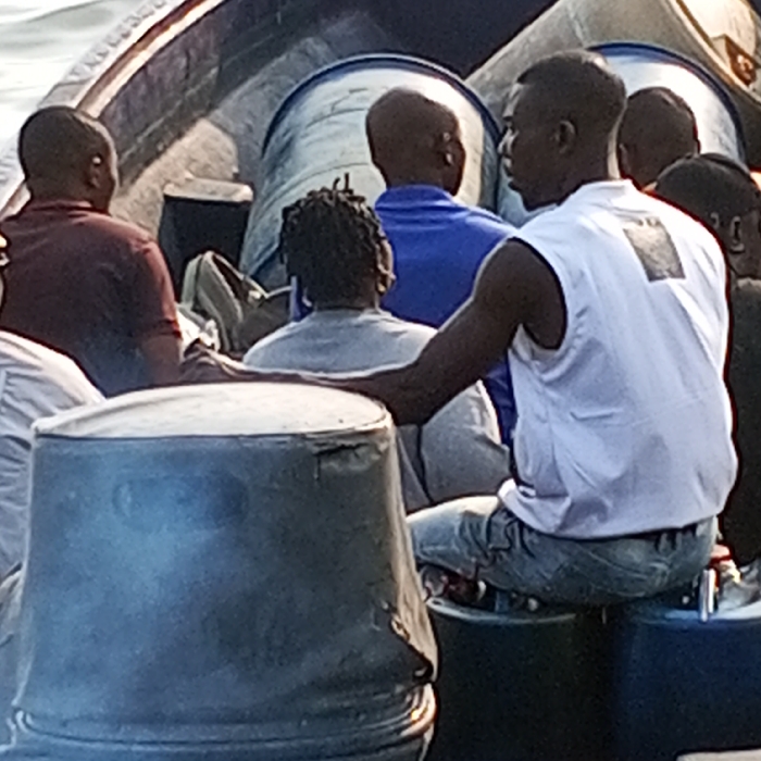 The image depicts at least seven individuals on a boat in Koko, Nigeria. In the foreground, a dark-skinned male wearing a white sleeveless shirt and light-colored jeans sits on a dark blue cylindrical container, his left arm resting on a weathered gray barrel to his left. He is positioned facing right, partially away from the camera. Behind him, other individuals are visible, mostly facing away: one person with dreadlocks in a light gray top, another in a blue short-sleeved shirt, and a person in a dark reddish-brown shirt. Multiple large, cylindrical barrels are present within the boat, including the gray barrel in the foreground, the dark blue barrel used as a seat, and several lighter-colored barrels with blue bands in the background. The boat is on a body of water, visible in the upper left, under daylight conditions.