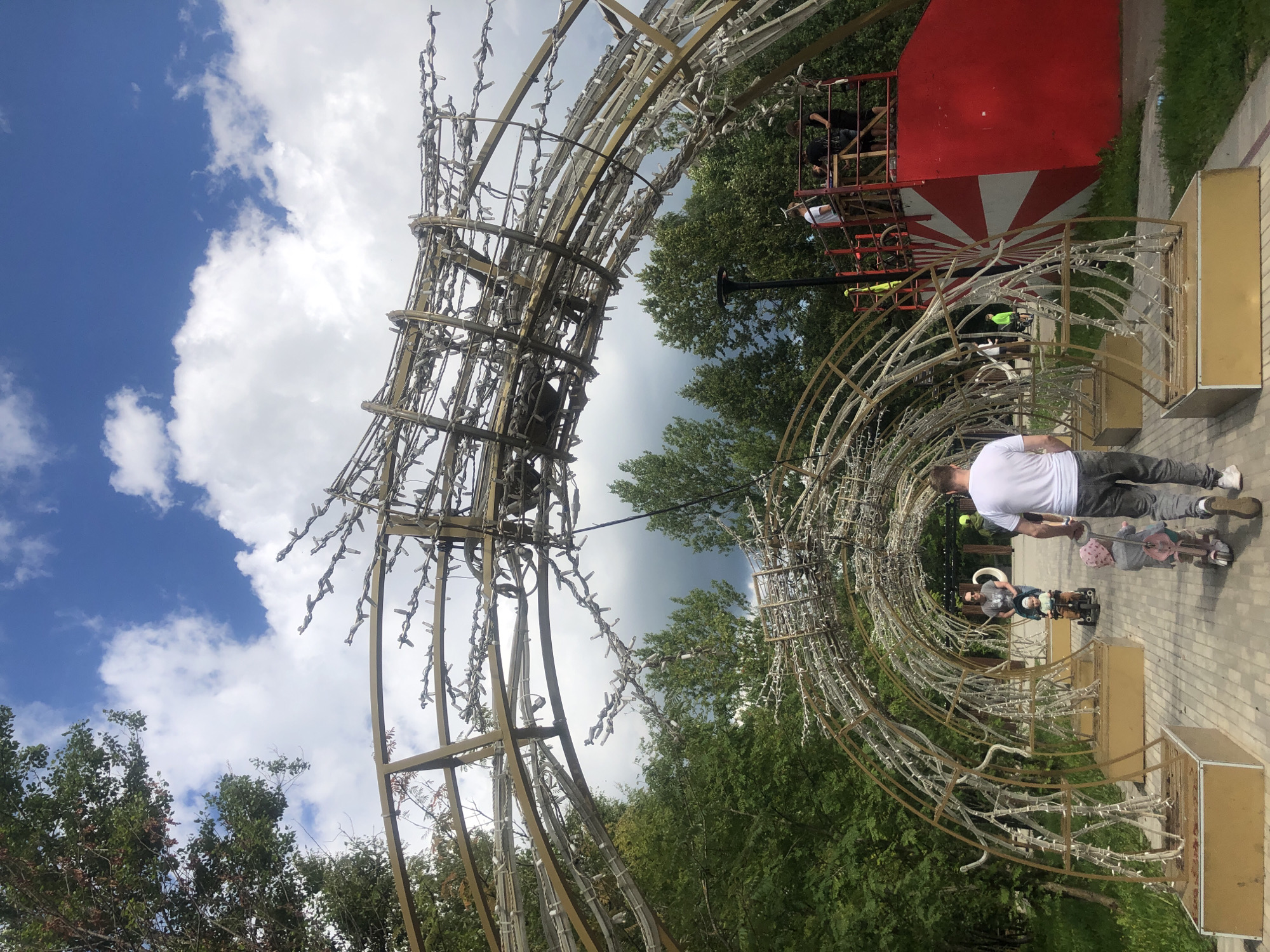 In Tutaev, Russia, an outdoor scene depicts a park pathway beneath a large, arching metal structure adorned with intricate lighting elements. The pathway is paved with light-colored bricks.
To the left of the pathway, lush green trees provide a natural backdrop. The sky above is a bright blue with scattered white clouds. The metal archway is constructed with a framework of curved beams and crisscrossing wires, upon which numerous small, white lights are attached, giving it a festive or decorative appearance.
Several people are present. In the foreground, a man in a white t-shirt and grey pants walks away from the camera, pushing a young child on a small tricycle. The child wears a pink hat and dress. Further down the path, a woman wearing sunglasses and a grey t-shirt is seen with a child on a similar tricycle. Another man in a white t-shirt is visible in the middle ground, also walking away from the camera.
In the background, a large, red structure with white radial stripes is partially visible on the right side. Above this structure, several individuals are seen on a raised platform, appearing to be working on or around the decorative archway. A lamppost is also present near the pathway. The overall atmosphere suggests a public park or event space, possibly during preparation for a celebration or installation.