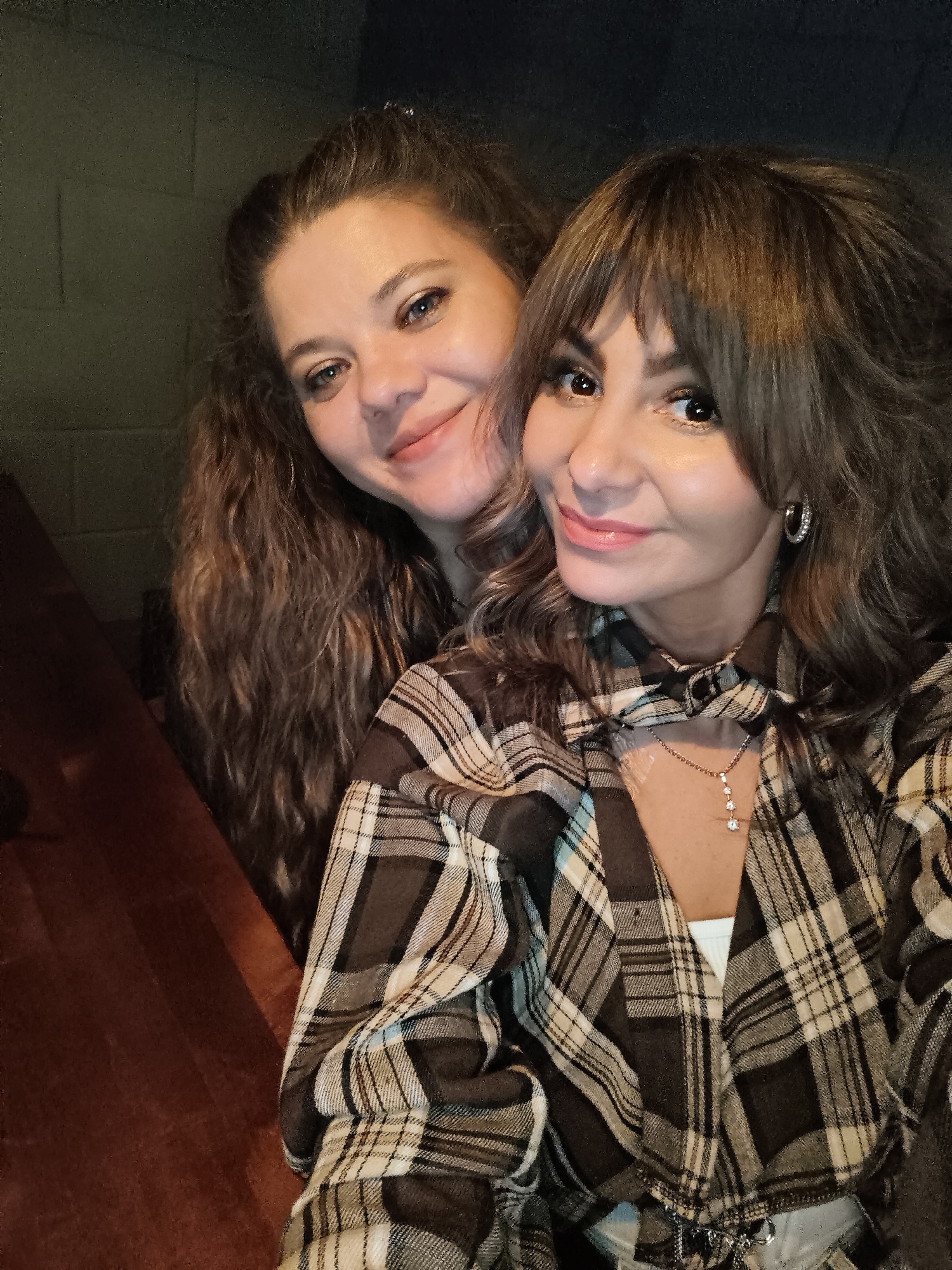 Two women are closely posed together, likely for a selfie, indoors. One woman has long, wavy brown hair and is standing behind the other woman, who is wearing a brown and beige plaid shirt. The woman in front has bangs and an elaborate necklace. There is dark, indistinct wall in the background. There is also a section of dark wood visible on the left side of the image. The location, Chaikovsky, Russia, is not discernible from the visual content of the image.